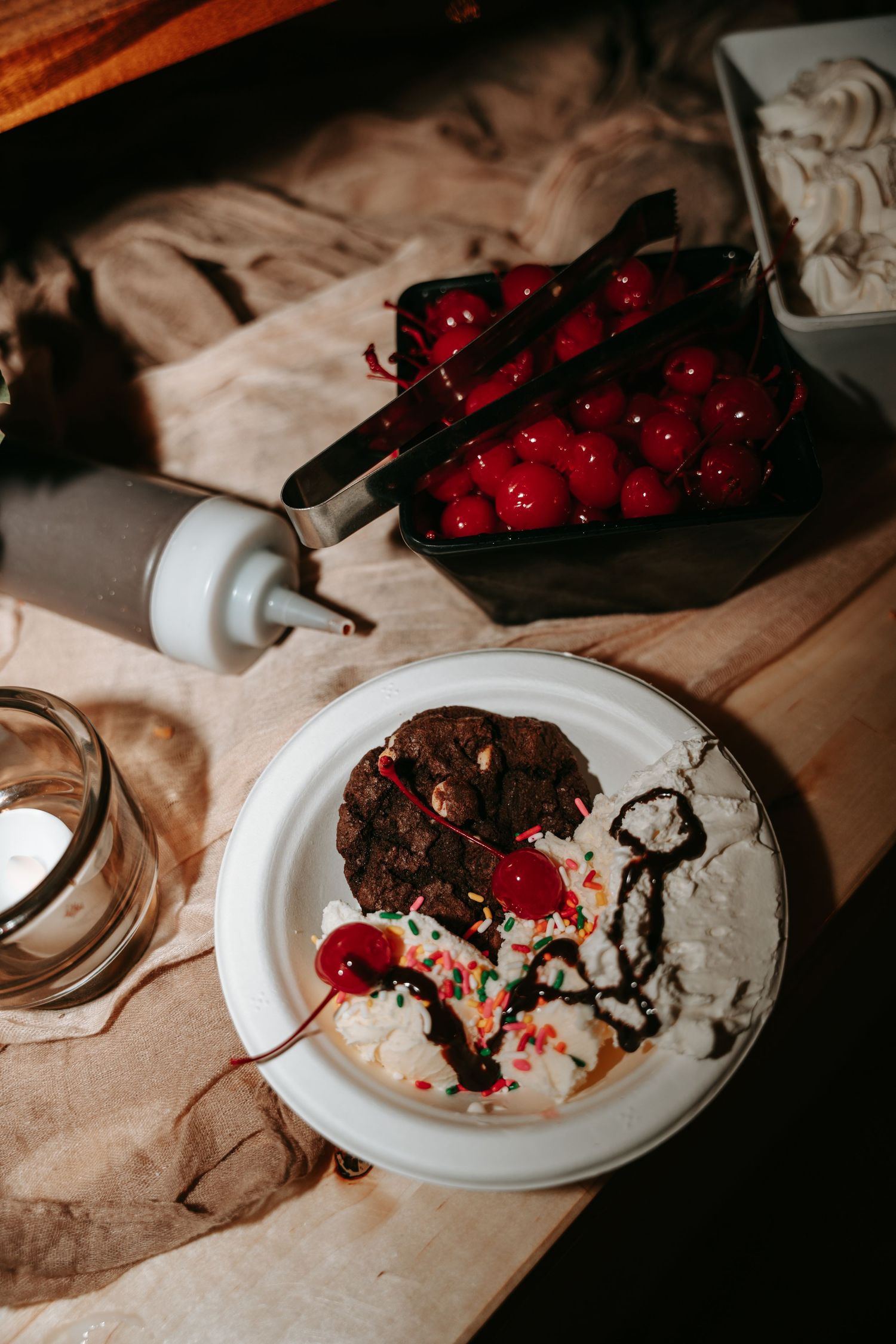 Chocolate brownie dessert plate with whipped cream, cherries, and sprinkles beside a tray of maraschino cherries on wood.