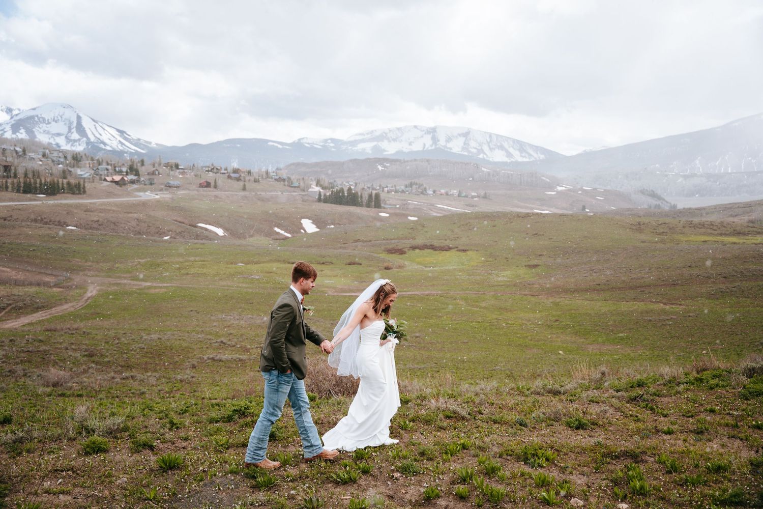 Groom helps bride with her veil while walking through a scenic green alpine meadow with mountains.