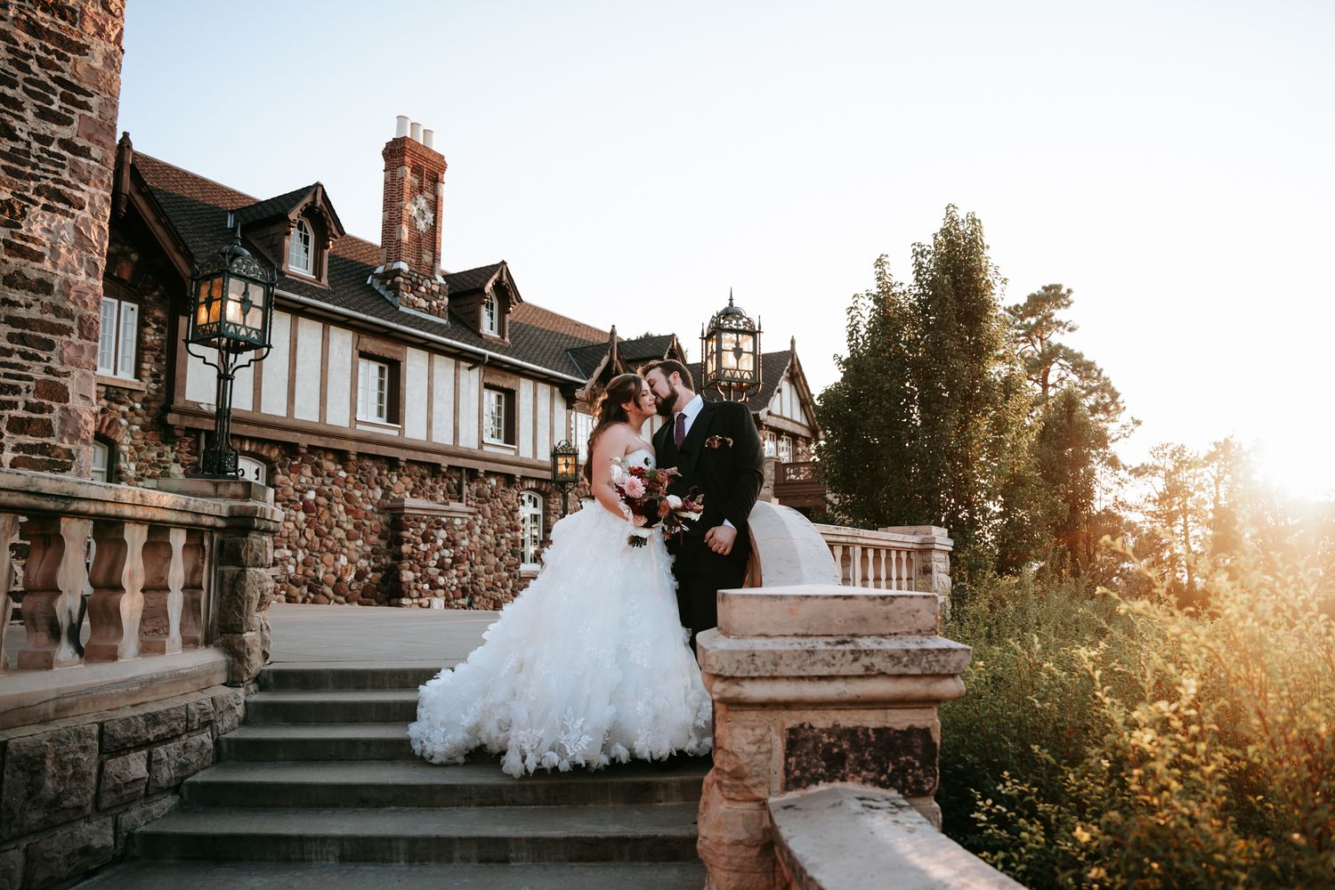 Bride and groom sharing a romantic kiss on stone steps outside a grand Tudor-style mansion at sunset.