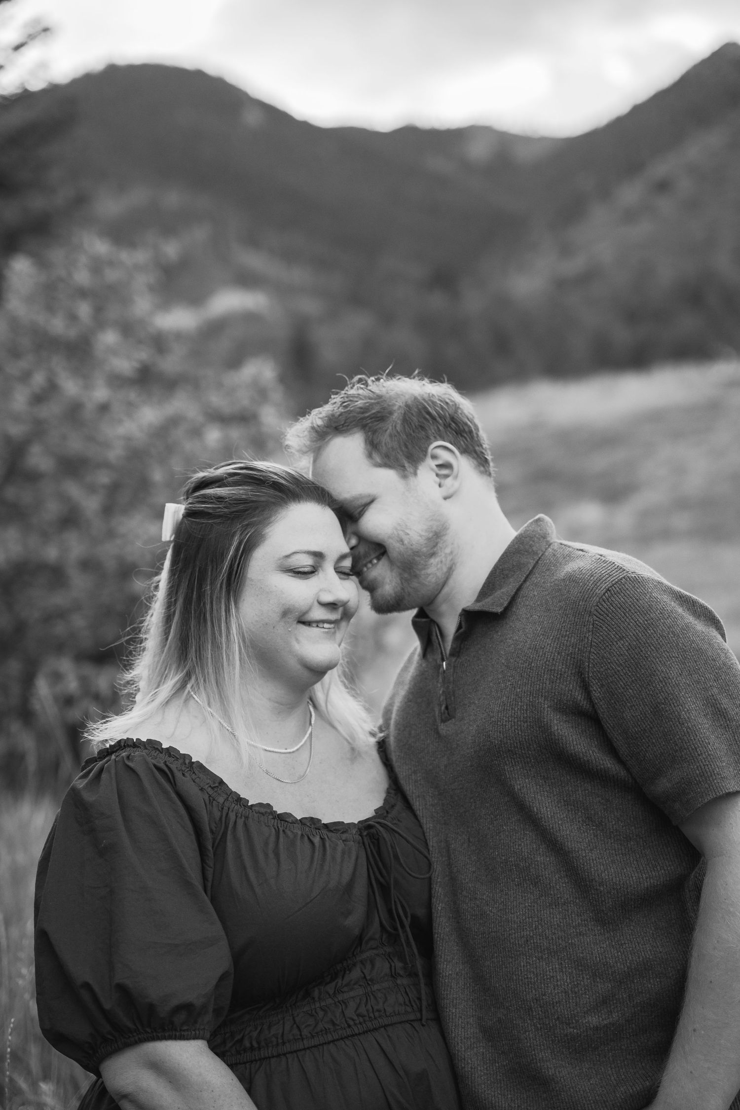 Black and white photo of a man kissing a smiling woman's temple in a mountain meadow.