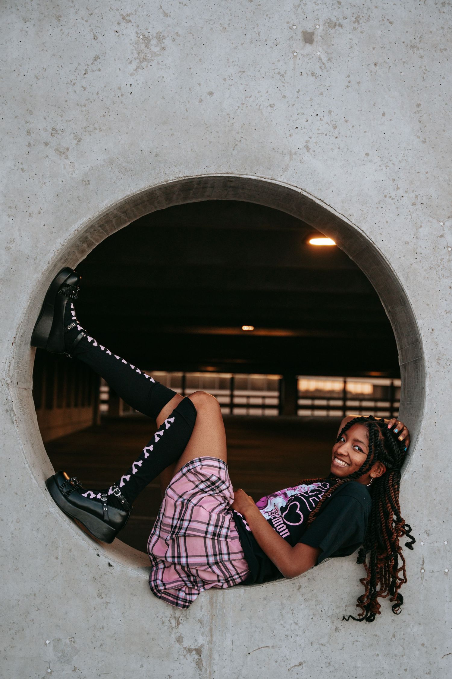 Young woman with braids posing in a circular concrete opening wearing plaid skirt and black combat boots.