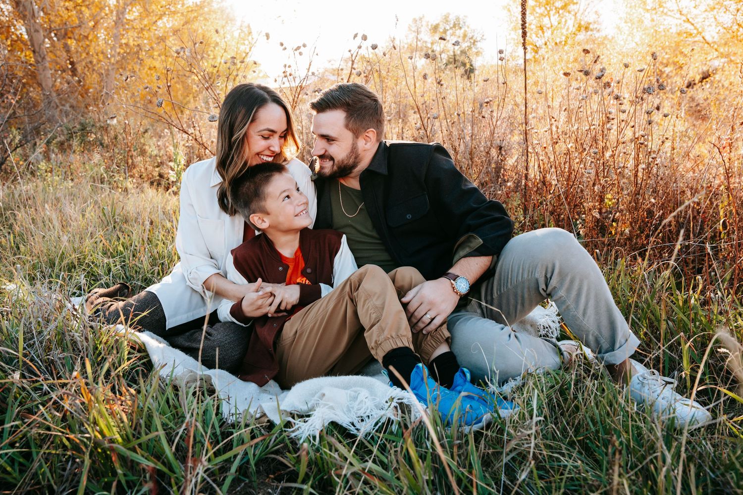 A happy family of three sits together in a golden autumn field, sharing laughter and warm embraces.