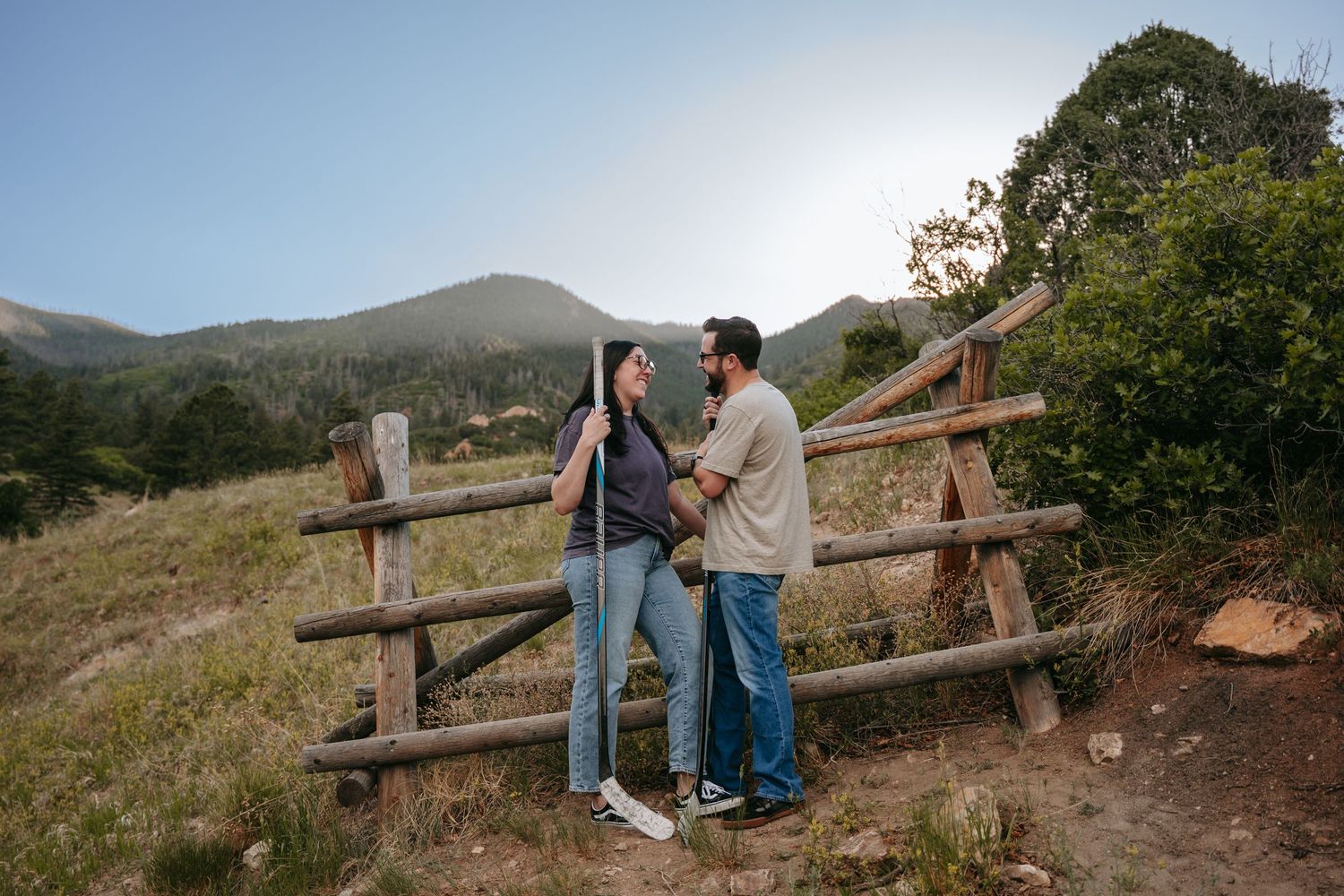 Couple stands by rustic wooden fence in mountain meadow, sharing a tender moment with scenic hills and trees in background.
