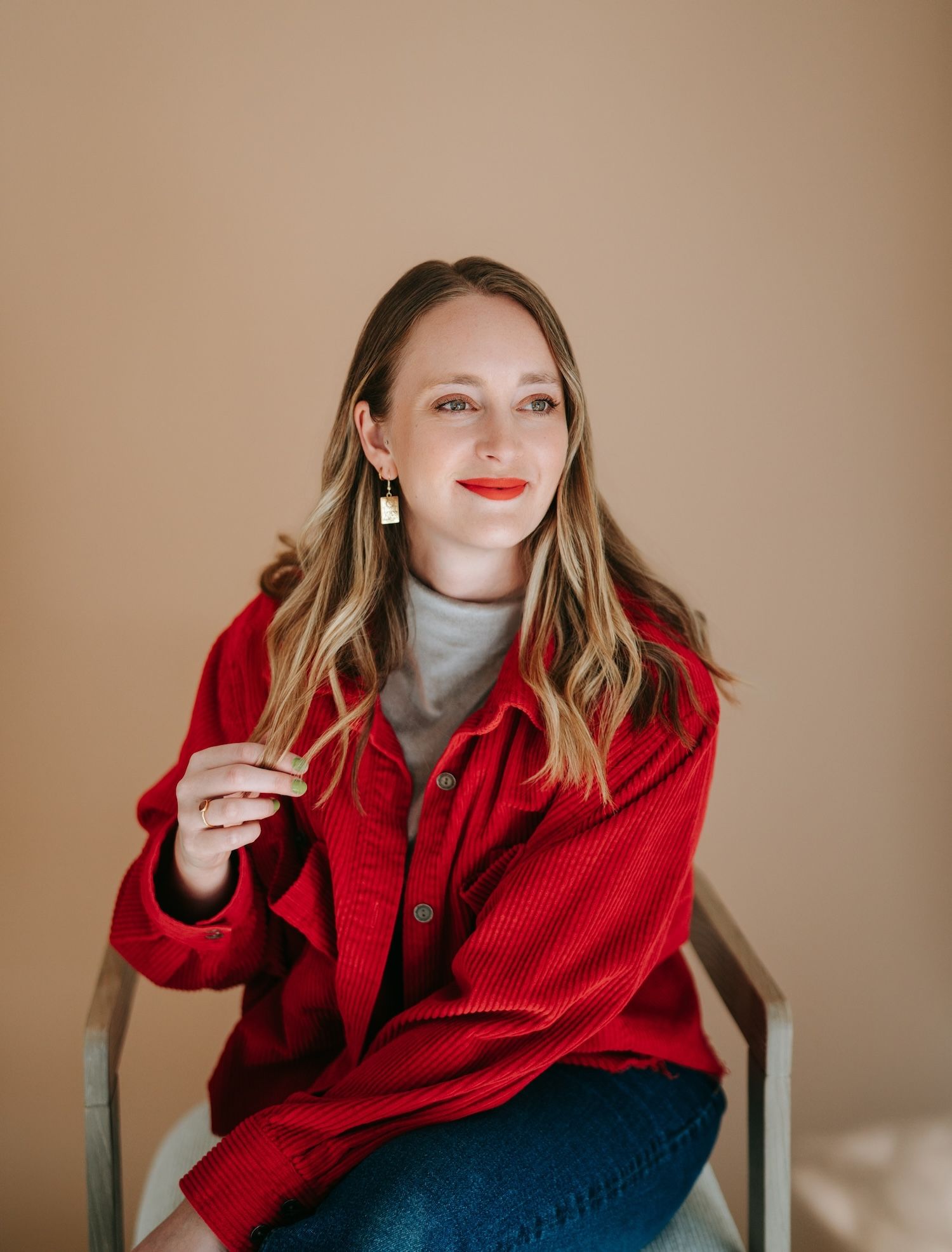 Smiling woman in red corduroy jacket sitting in chair against beige background.