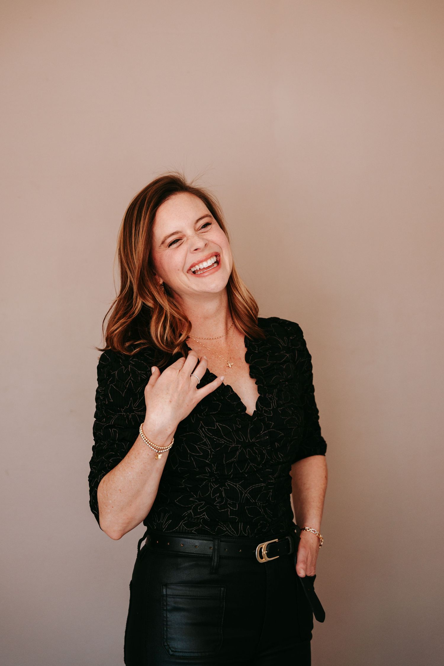 Laughing woman in black floral top and leather pants posing against neutral beige background indoors.