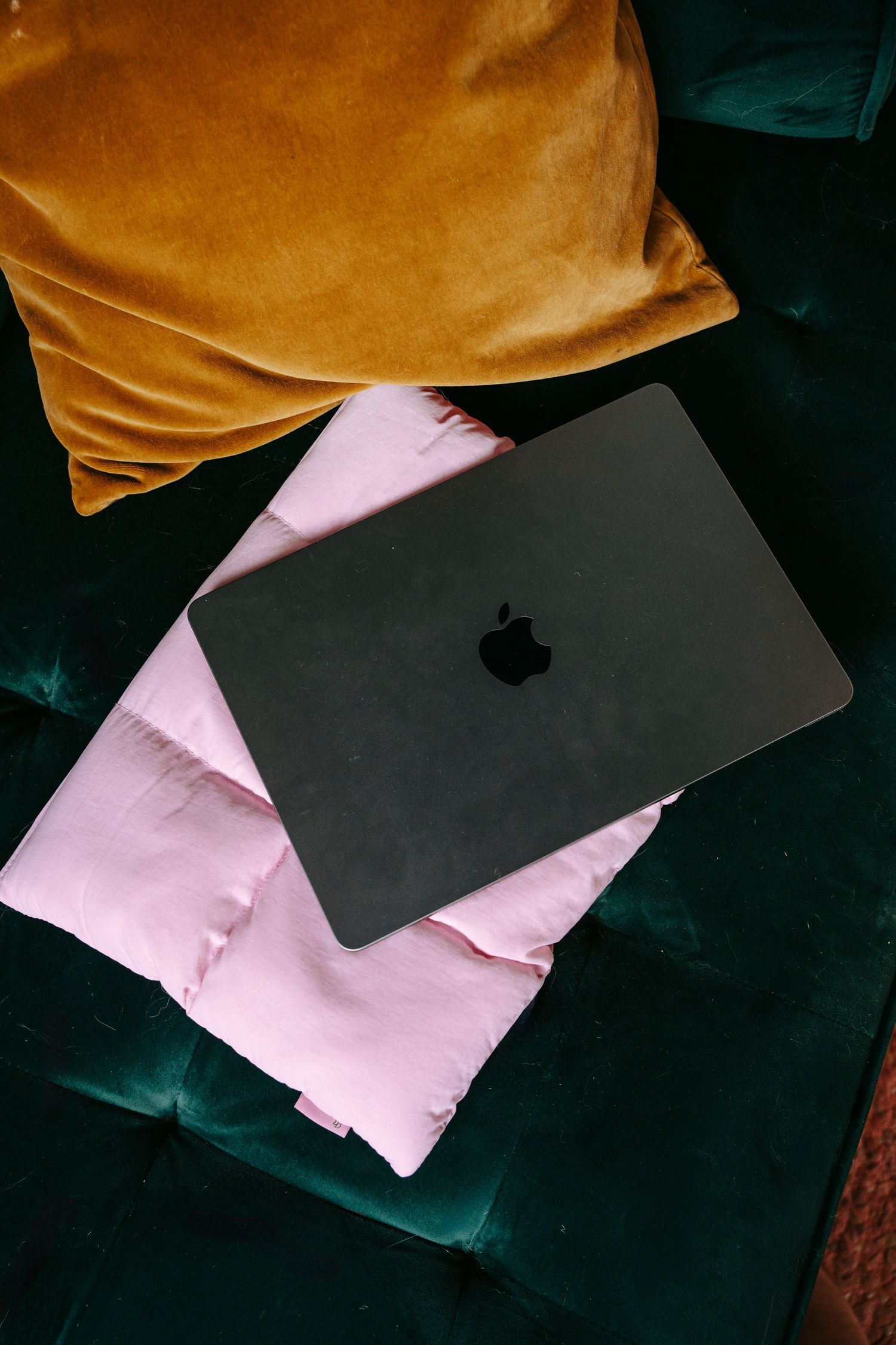 Dark gray Apple MacBook laptop resting on pink and gold pillows on a green velvet couch.