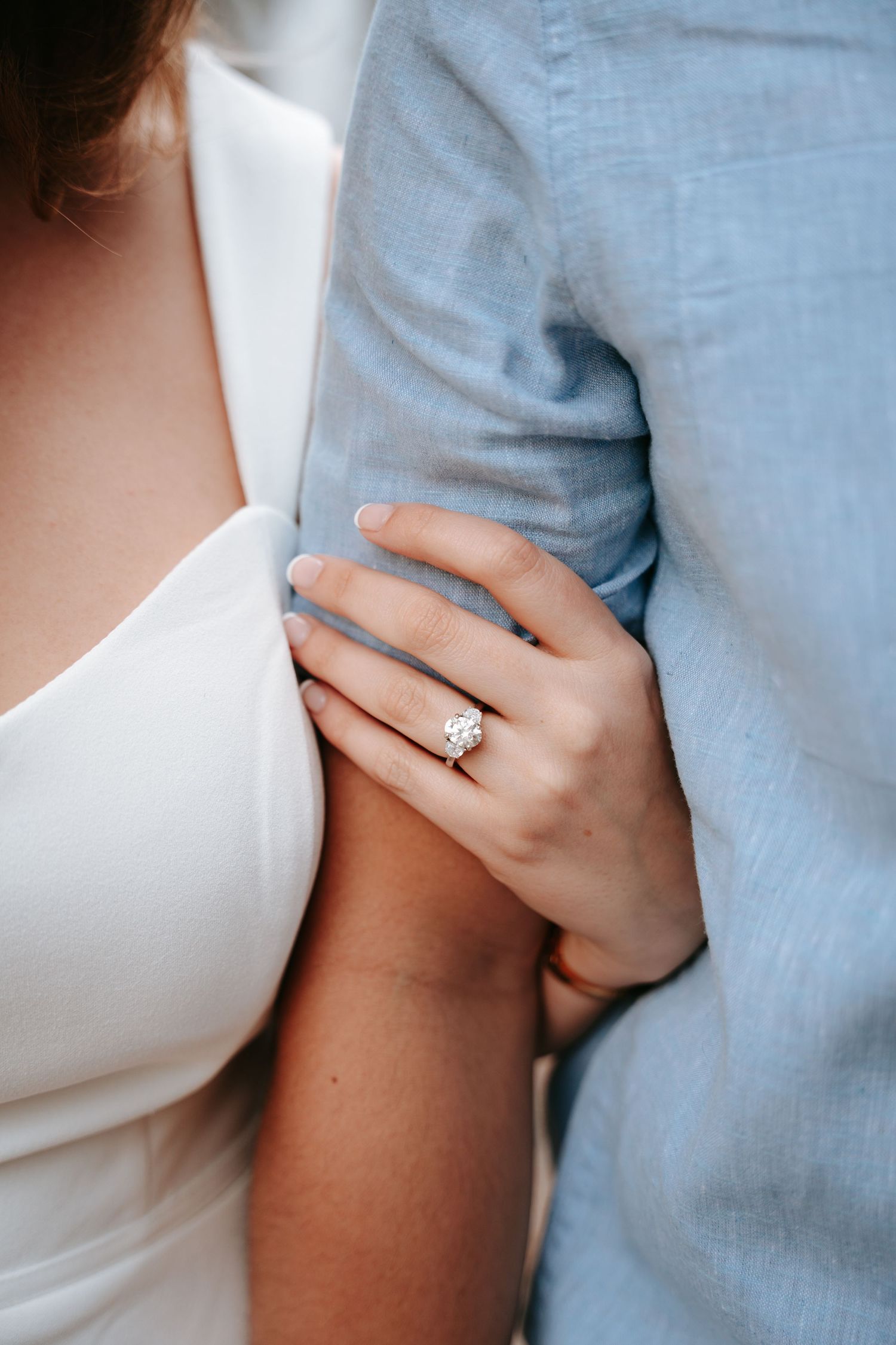 Close-up of a woman's hand wearing a diamond engagement ring while holding a man's arm in a blue shirt.
