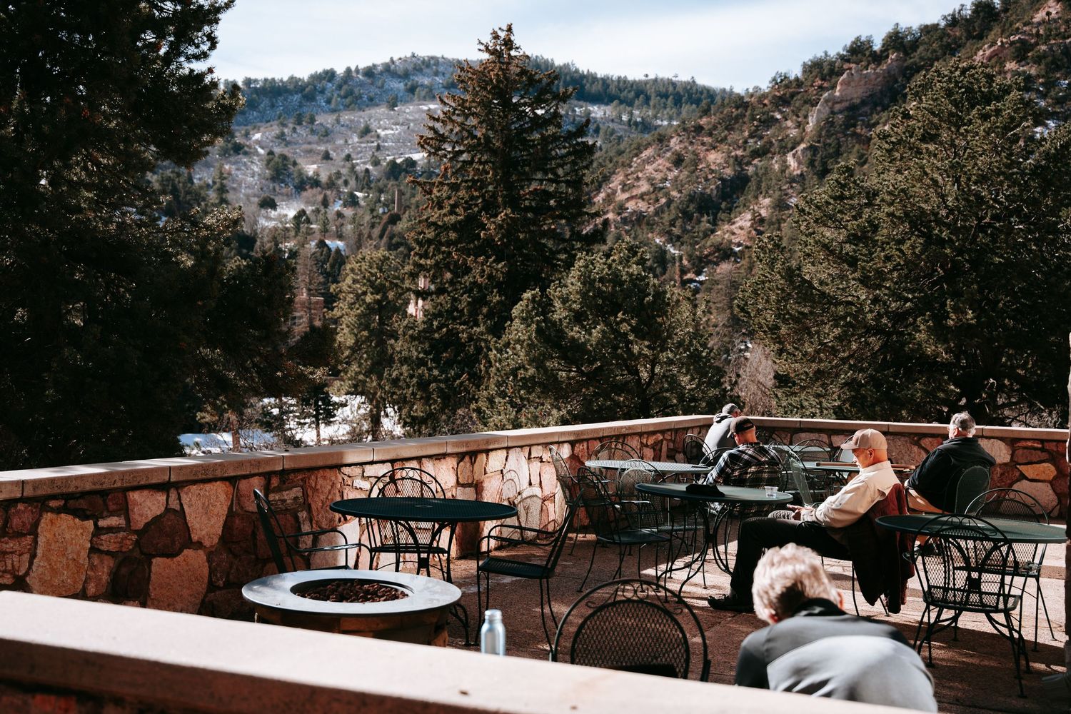 Guests dine at tables on a stone-walled mountain terrace overlooking evergreen trees and rocky hillsides under a pale blue sky.