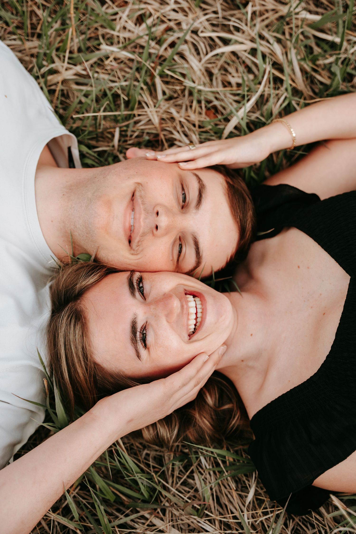 Smiling couple lying in grass with heads together, sharing a joyful moment outdoors.