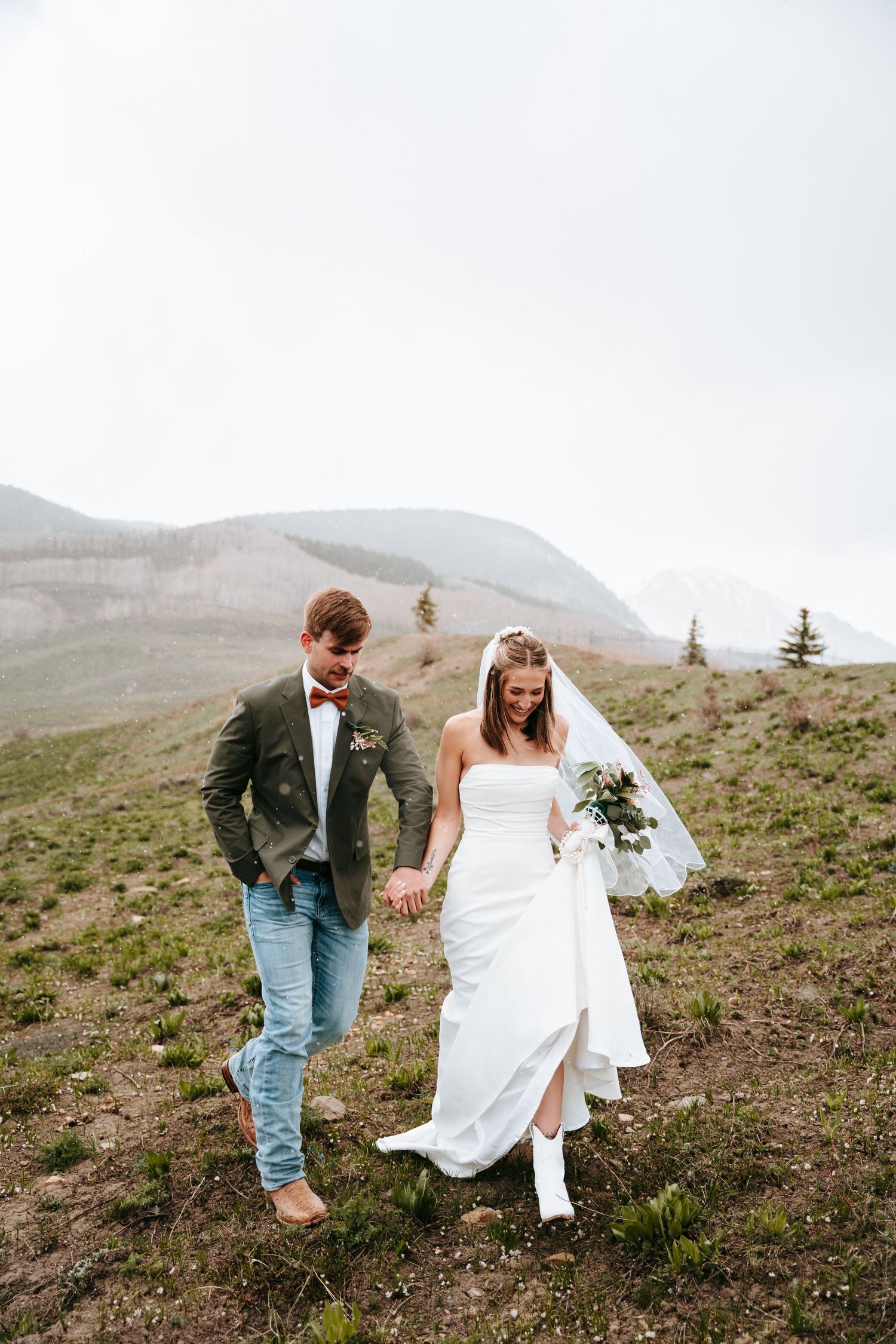 Bride in white dress and cowboy boots walking with groom in green jacket through mountain meadow.