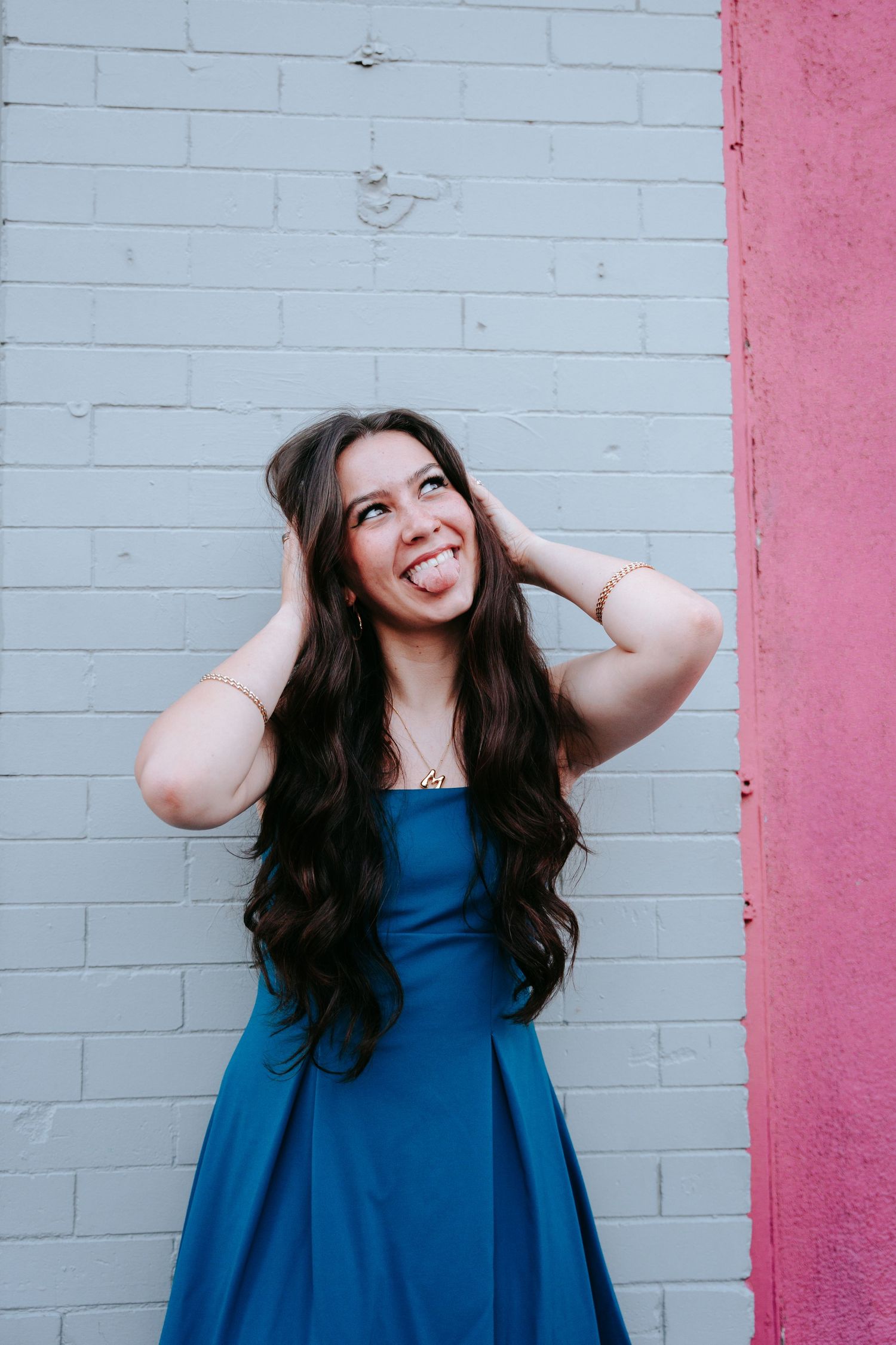 Joyful woman in blue dress sticking out tongue against white brick wall.