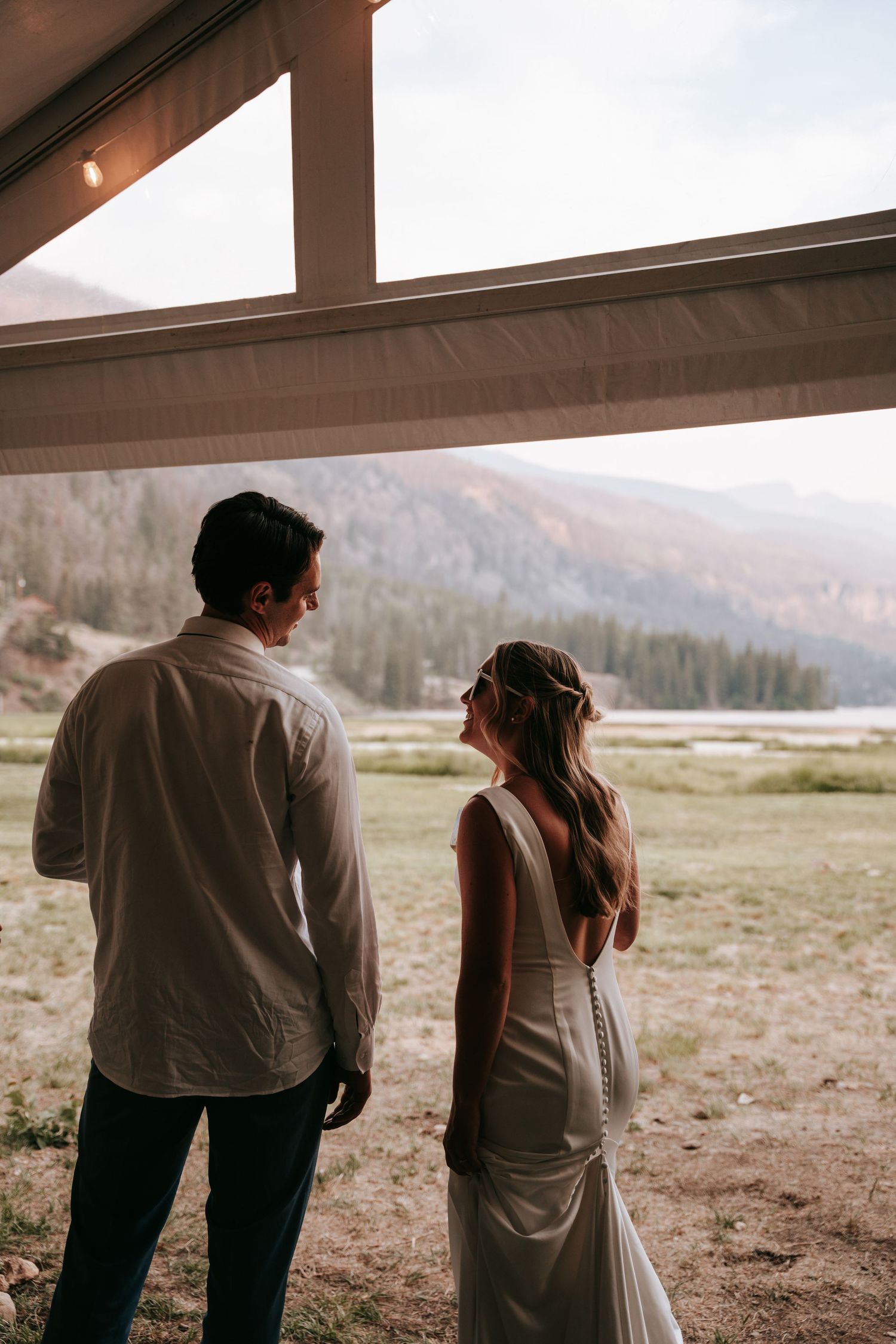 Bride in backless white gown and groom in white shirt admire mountain valley views at outdoor wedding venue.