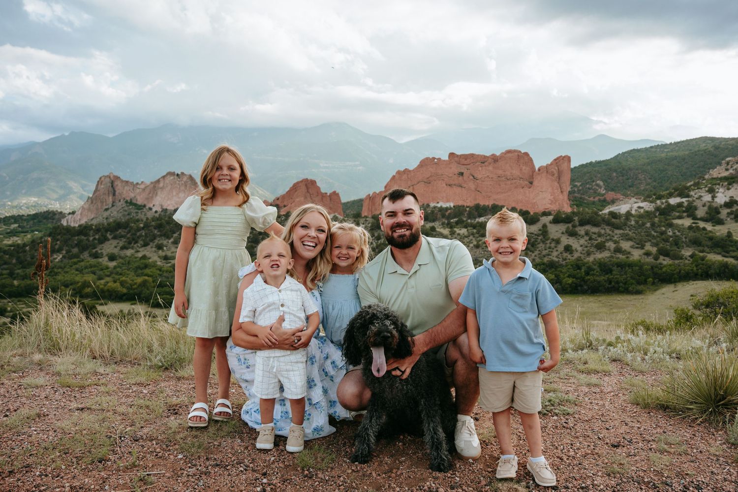 Family of five with a dog posing at Garden of the Gods, Colorado, with red rock formations and mountains in background.