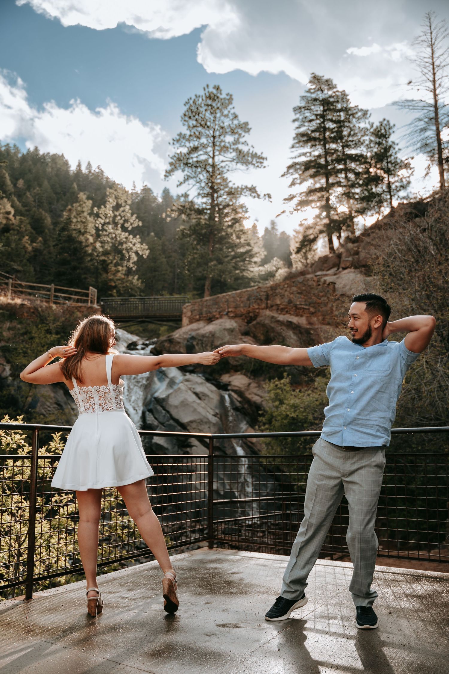 Couple dancing on a scenic overlook with a waterfall and tall trees in the background on a sunny day.