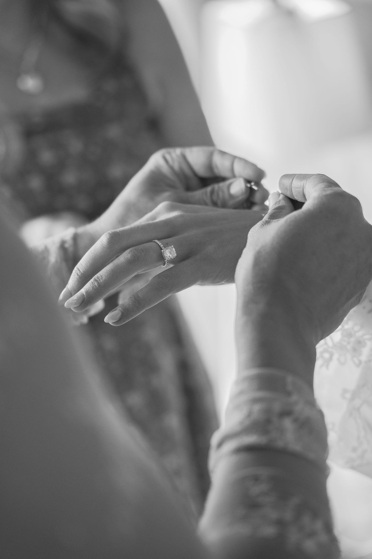 Black and white photo of hands fastening bride's wedding dress with diamond engagement ring visible.