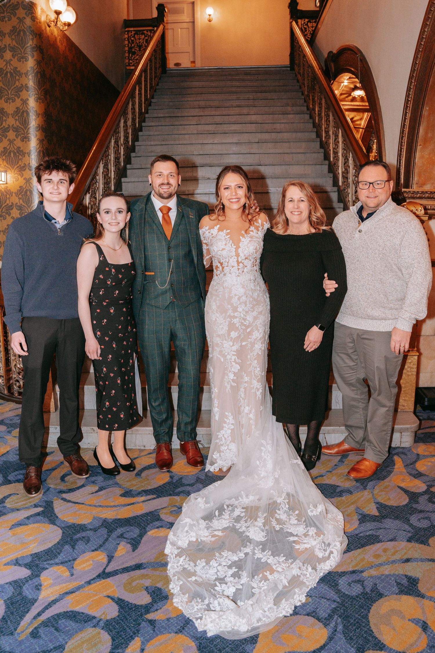 Wedding party poses on grand staircase with bride in lace gown and groom in green suit.