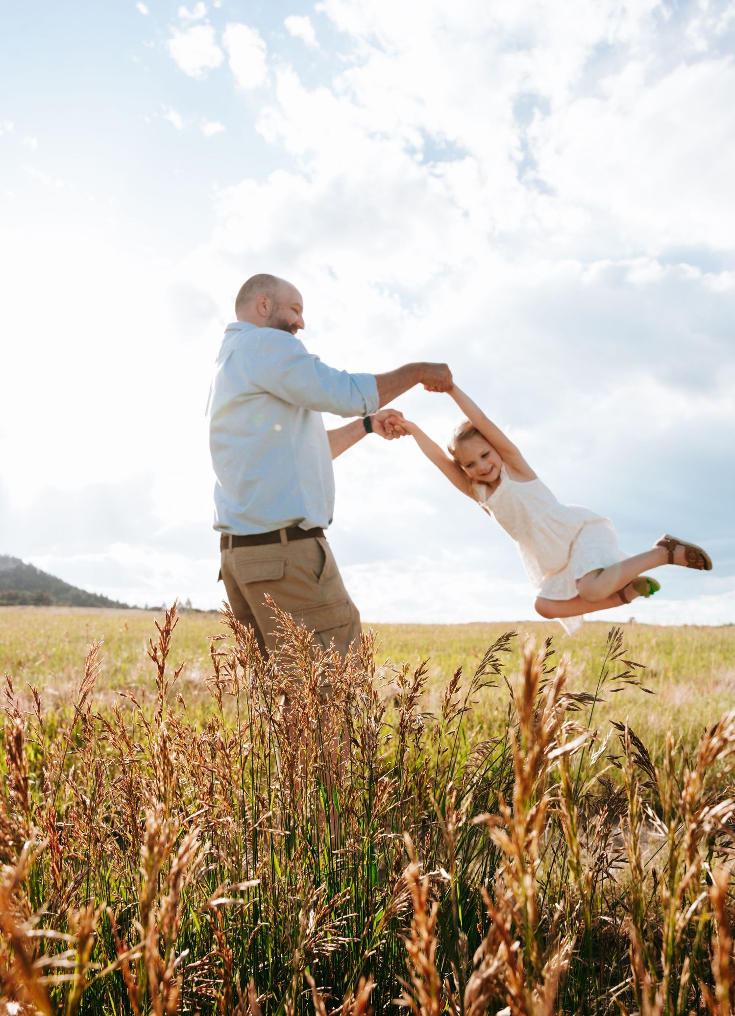 Father swinging young daughter by her hands in golden grass meadow under bright blue cloudy sky.