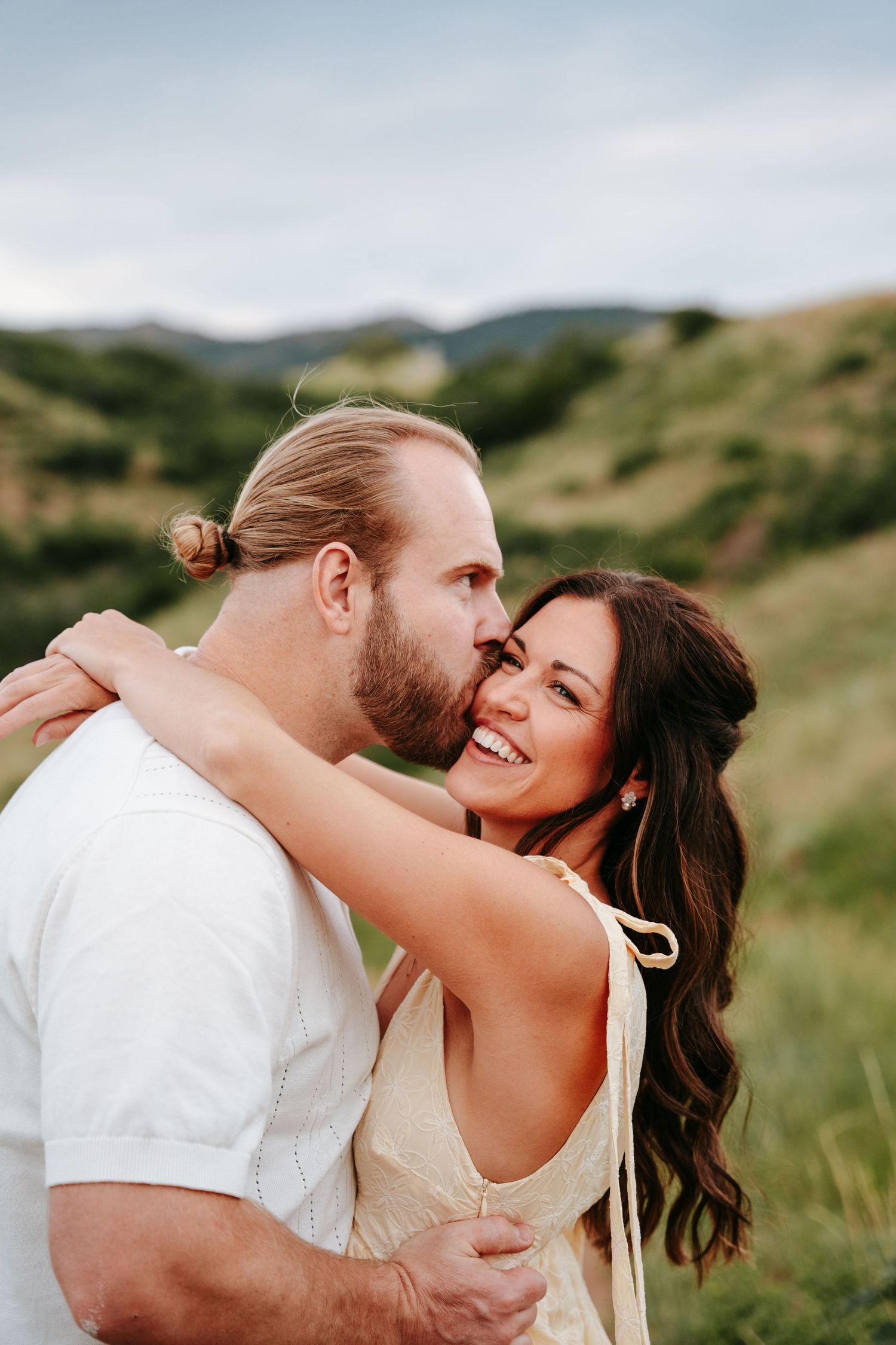 Woman smiling joyfully as man kisses her cheek in a green mountain landscape.