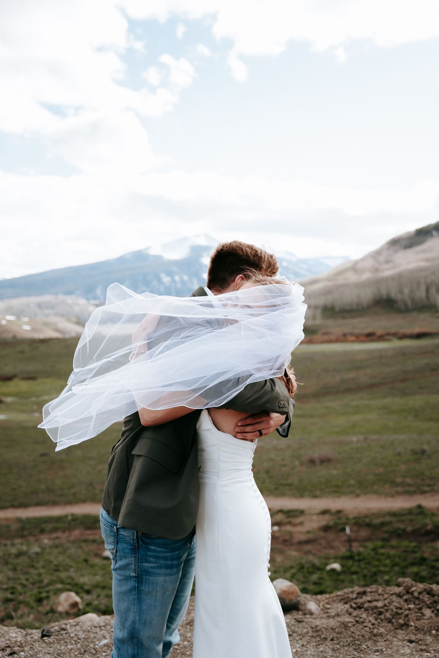Bride and groom share a romantic embrace as her veil blows in the wind against a mountain backdrop.