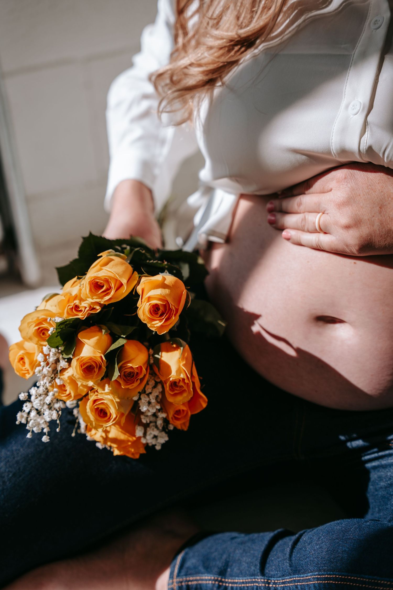 Pregnant woman in white shirt holding bouquet of orange roses while cradling baby bump.