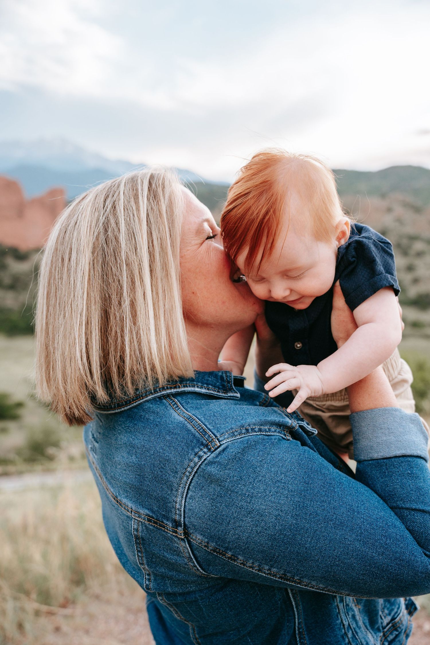 Blonde woman kissing a happy red-haired baby outdoors with mountains behind.
