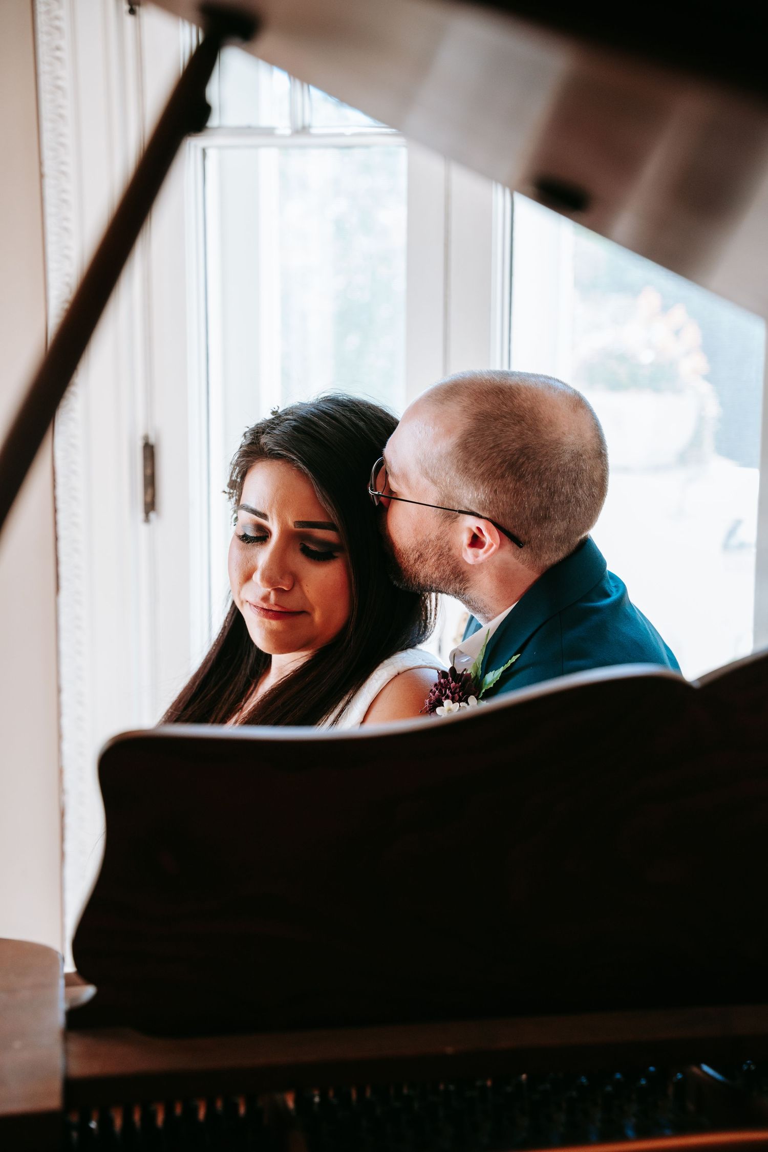 Bride and groom share an intimate moment together at a grand piano by a bright window.