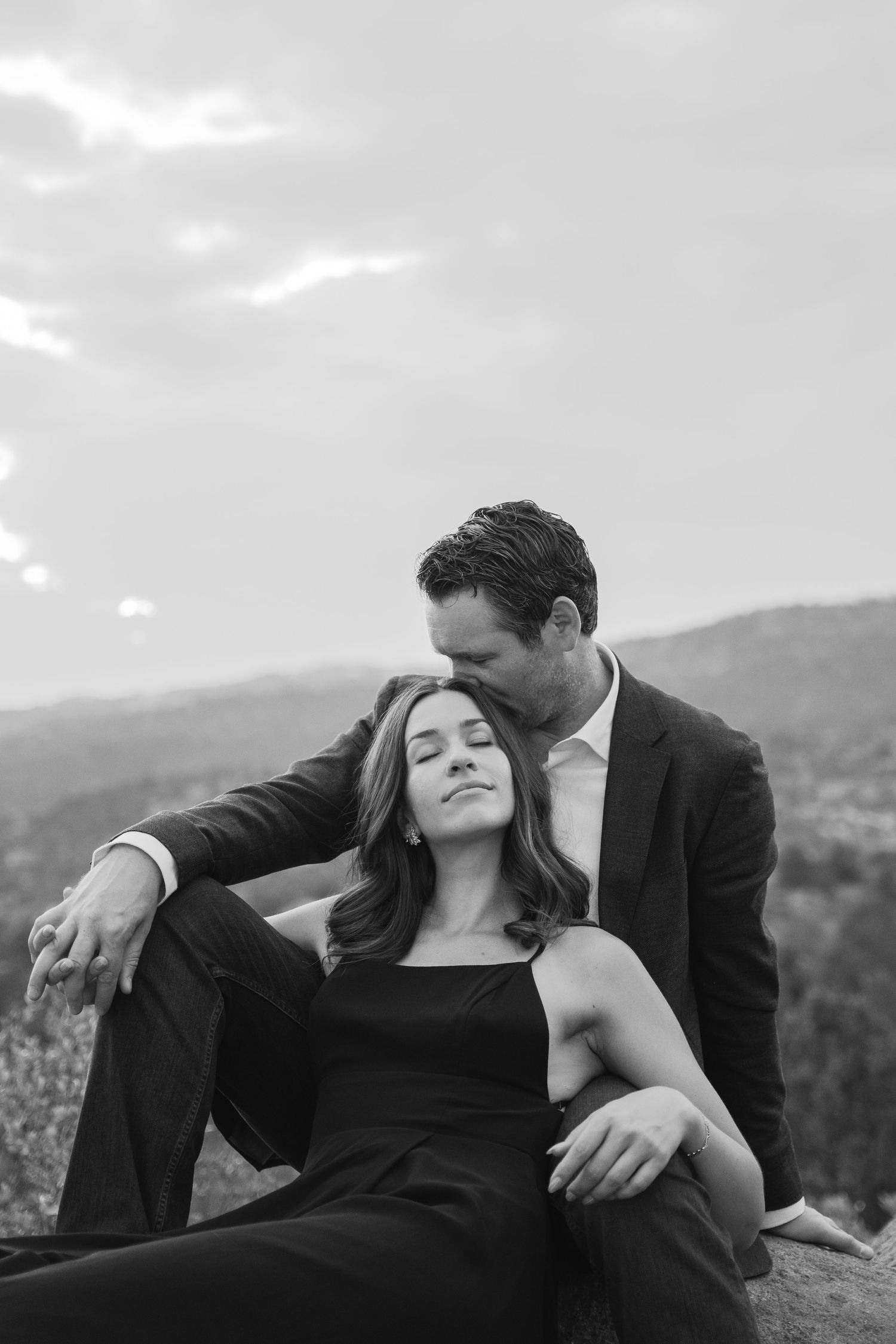 Black and white photo of a couple in elegant attire, man kissing woman's head outdoors with mountain views.