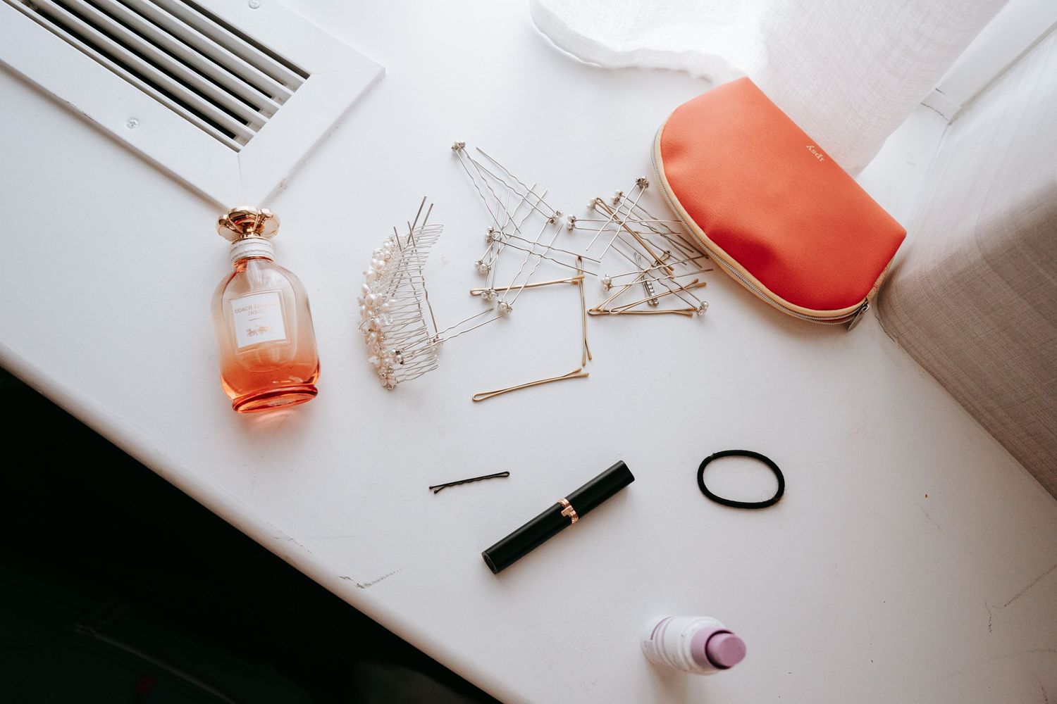 Bridal getting ready flat lay with rose perfume, hair pins, mascara, and coral makeup bag.