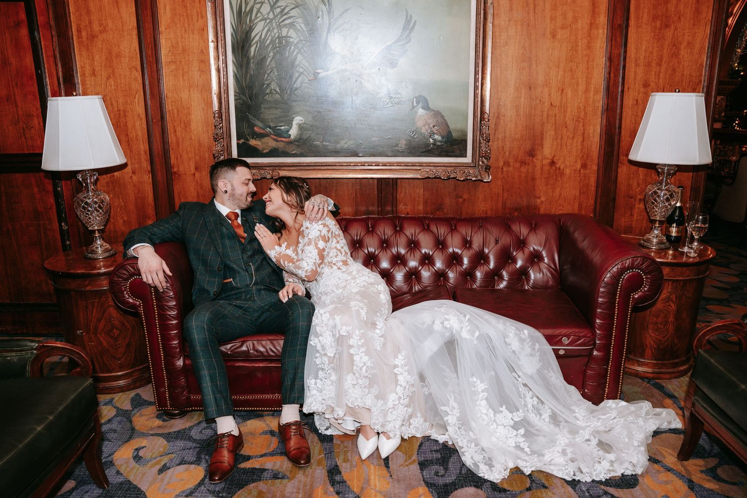 Bride and groom share an intimate moment on a tufted leather sofa beneath a classic oil painting.