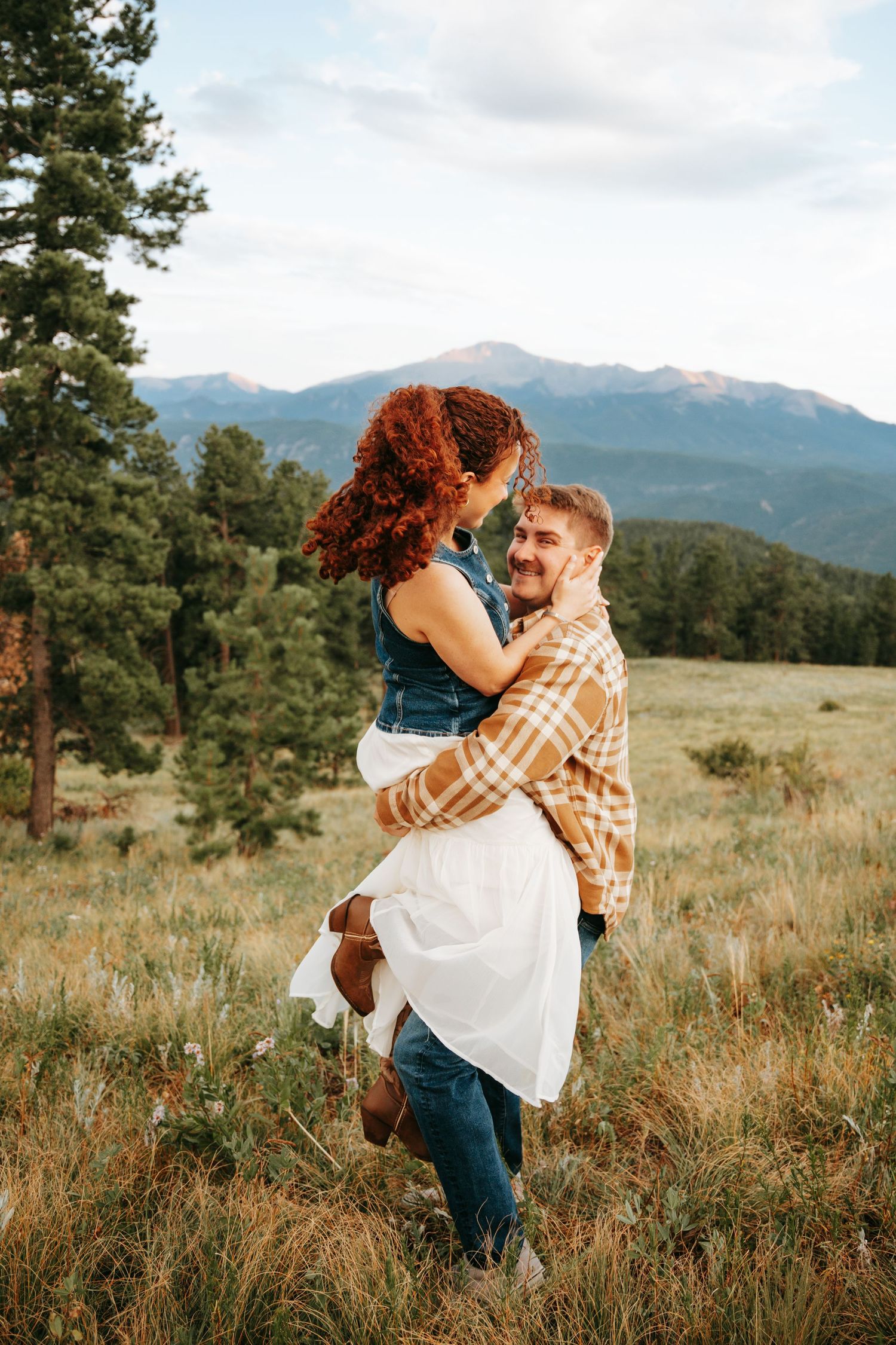Man in plaid shirt joyfully lifting curly red-haired woman in mountain meadow with scenic peak backdrop.