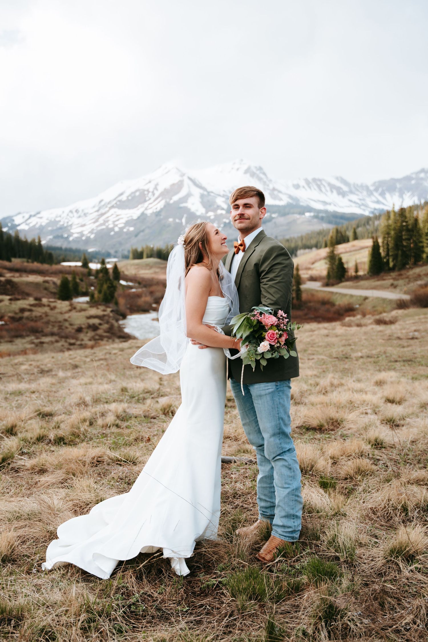 Groom holds pink rose bouquet while bride leans close in a golden meadow with snowy mountain peak.