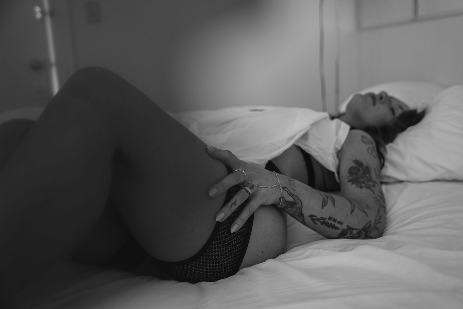 Black and white photo of a tattooed woman resting on a white bed in lingerie.
