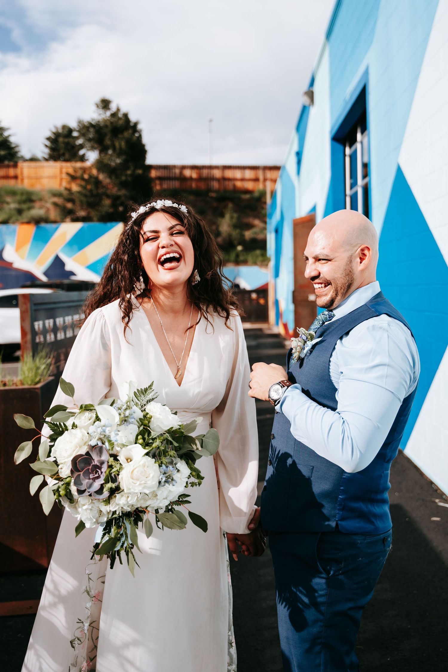 Laughing bride holding white bouquet and groom in blue vest pose outside a colorful mural wall.
