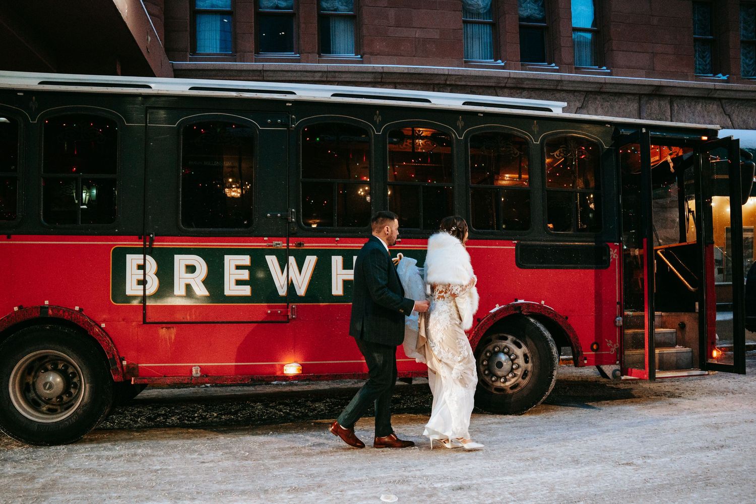 Bride and groom walking hand in hand in front of a red Brewhouse trolley in winter snow.