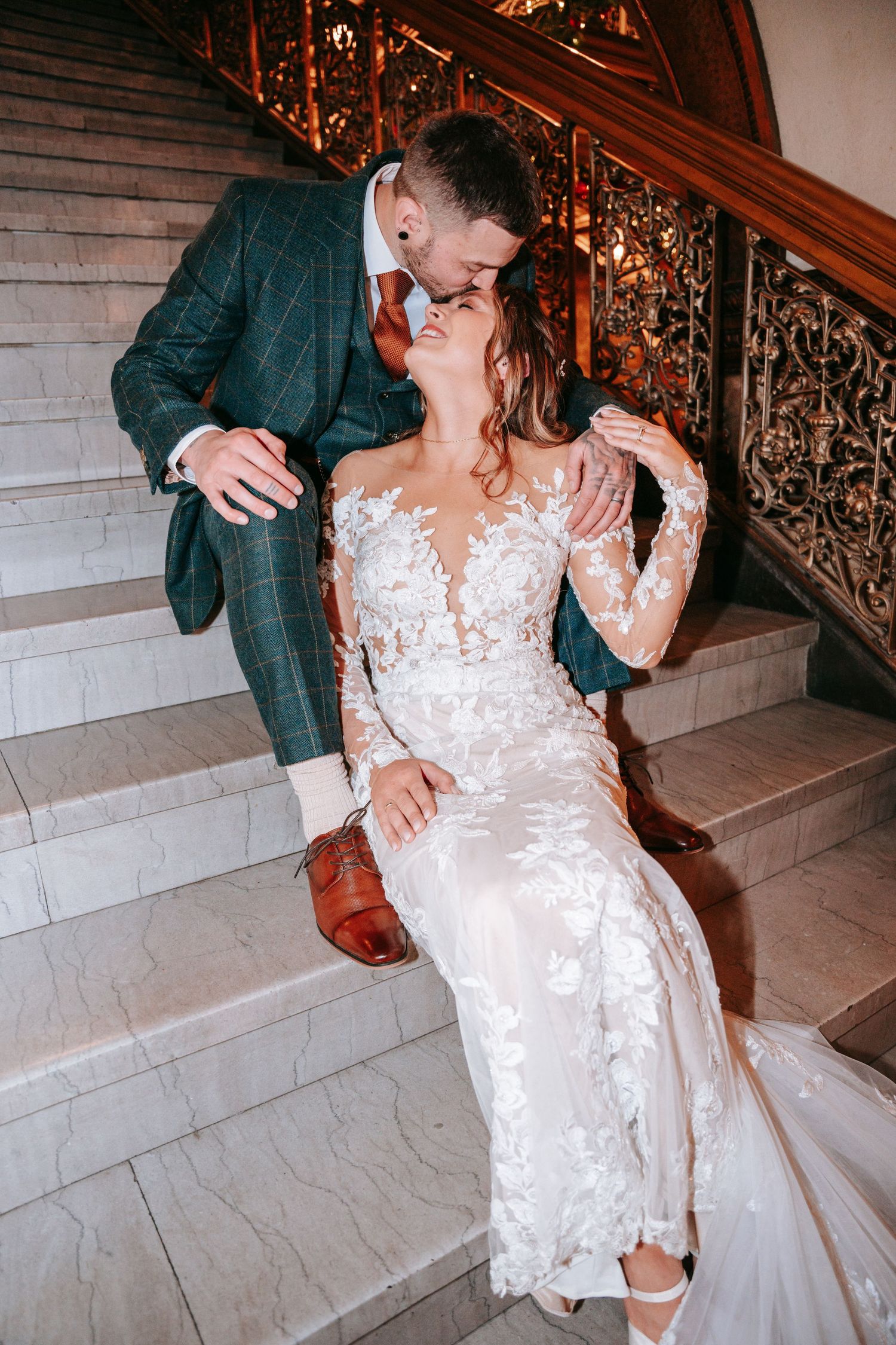 Bride in lace gown and groom in green plaid suit share a kiss on elegant marble staircase.