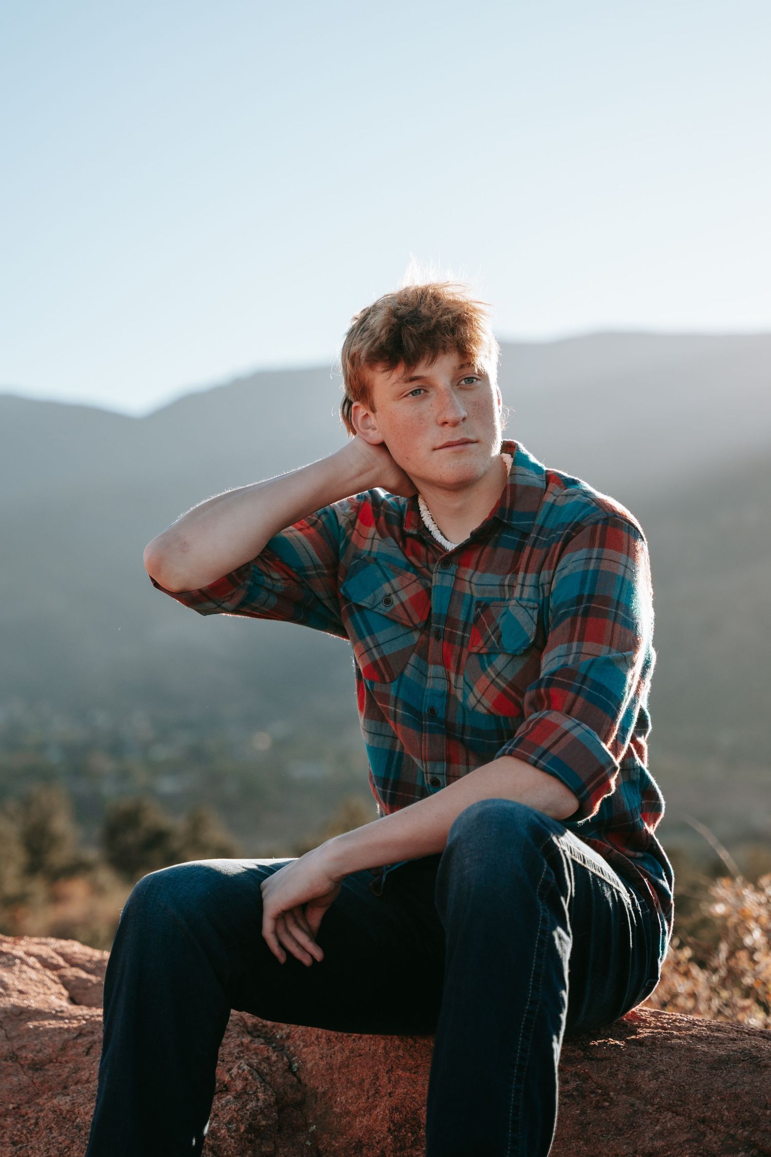 Young man in plaid flannel shirt sitting on red rocks with mountain landscape backdrop in golden hour light.