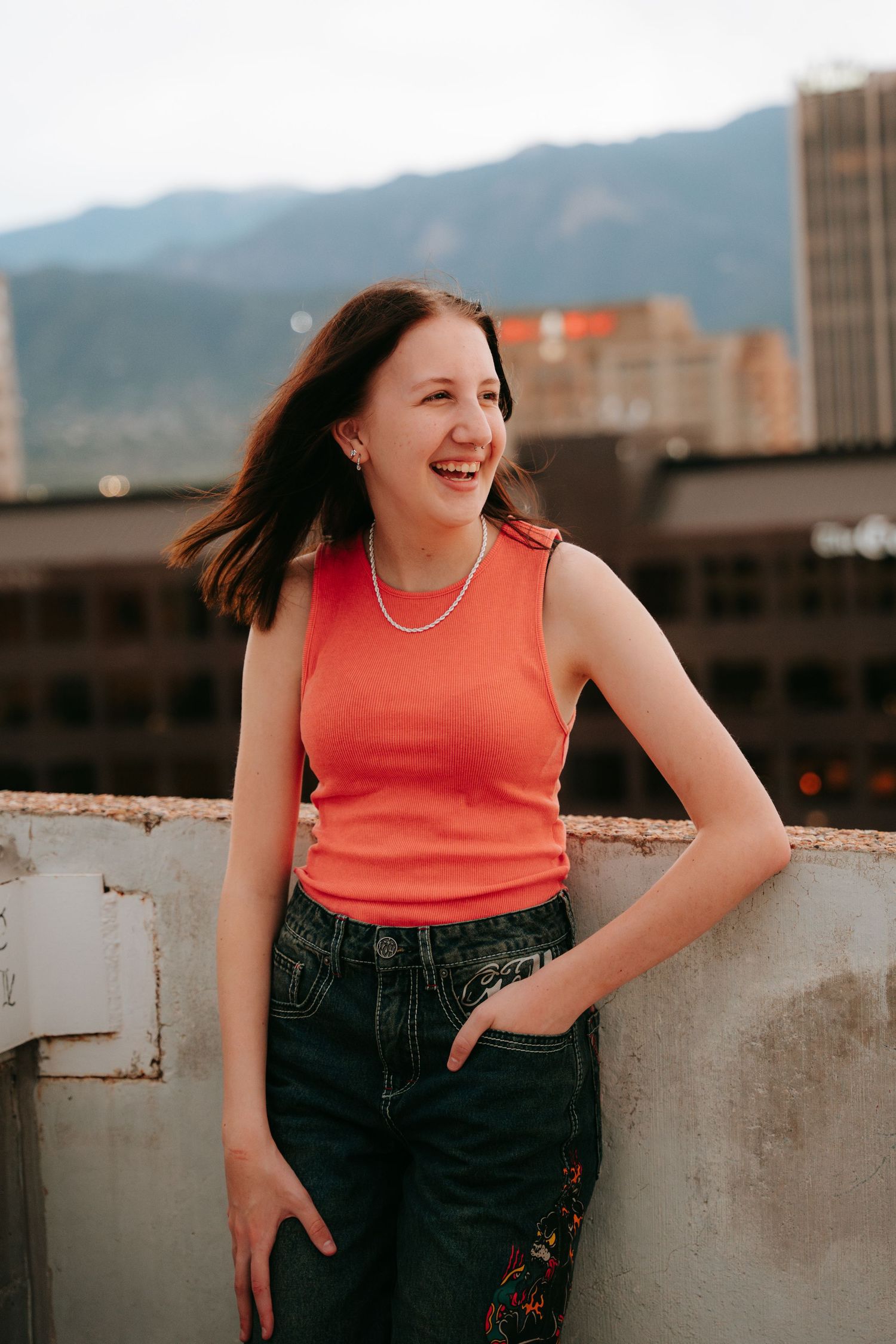 Happy young woman in orange tank top laughing on rooftop with mountain view.
