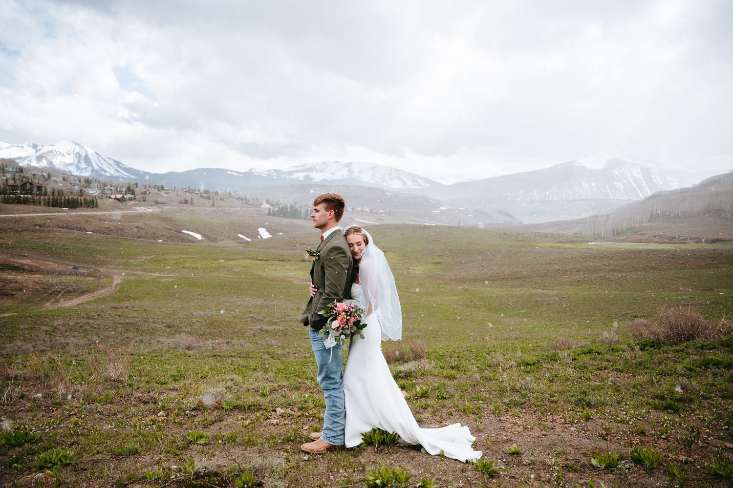 Bride and groom pose back-to-back in a vast green mountain meadow with snow-dusted peaks behind them.