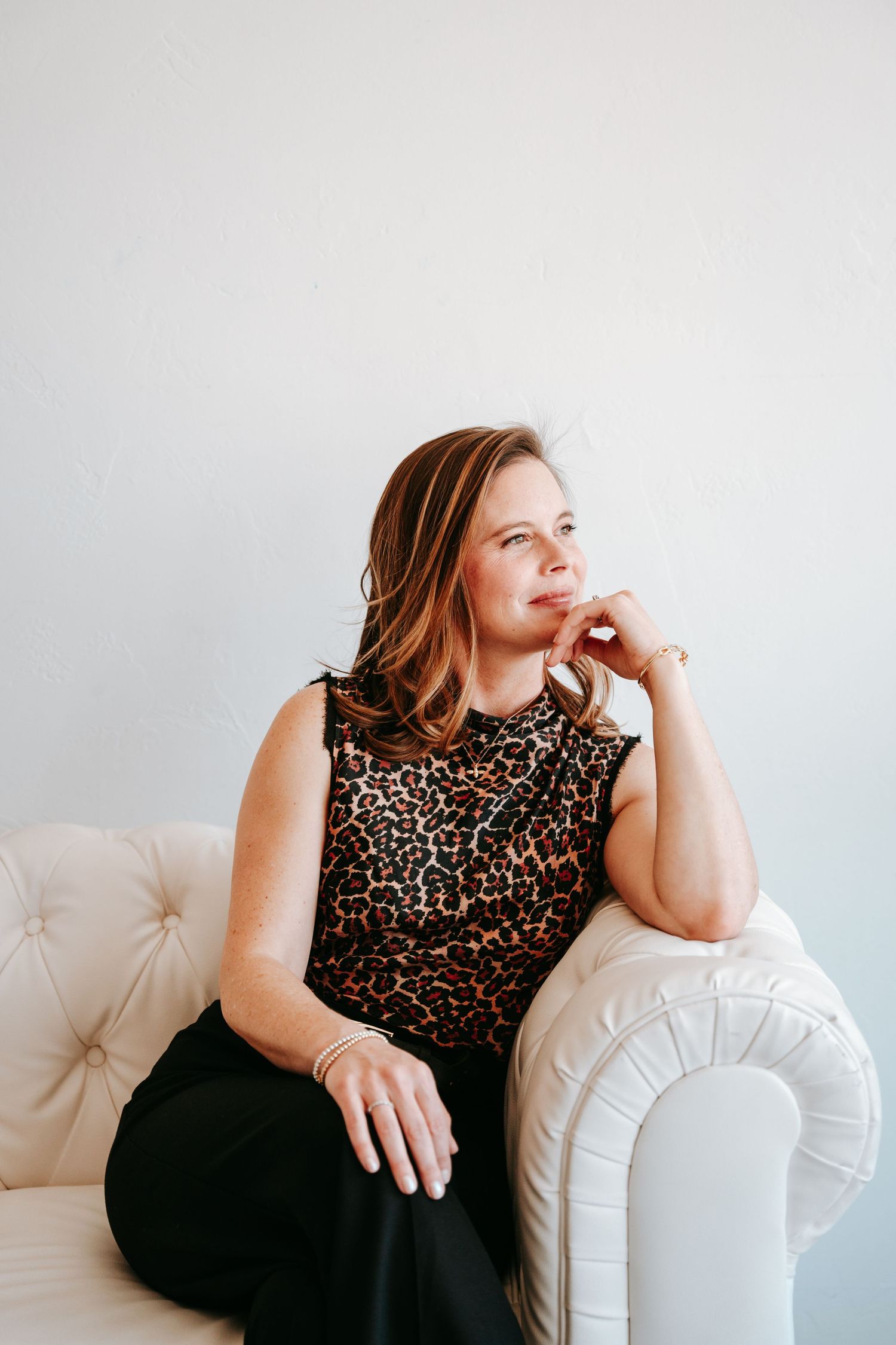 Woman in leopard print sleeveless top sitting thoughtfully in white tufted armchair against light wall.