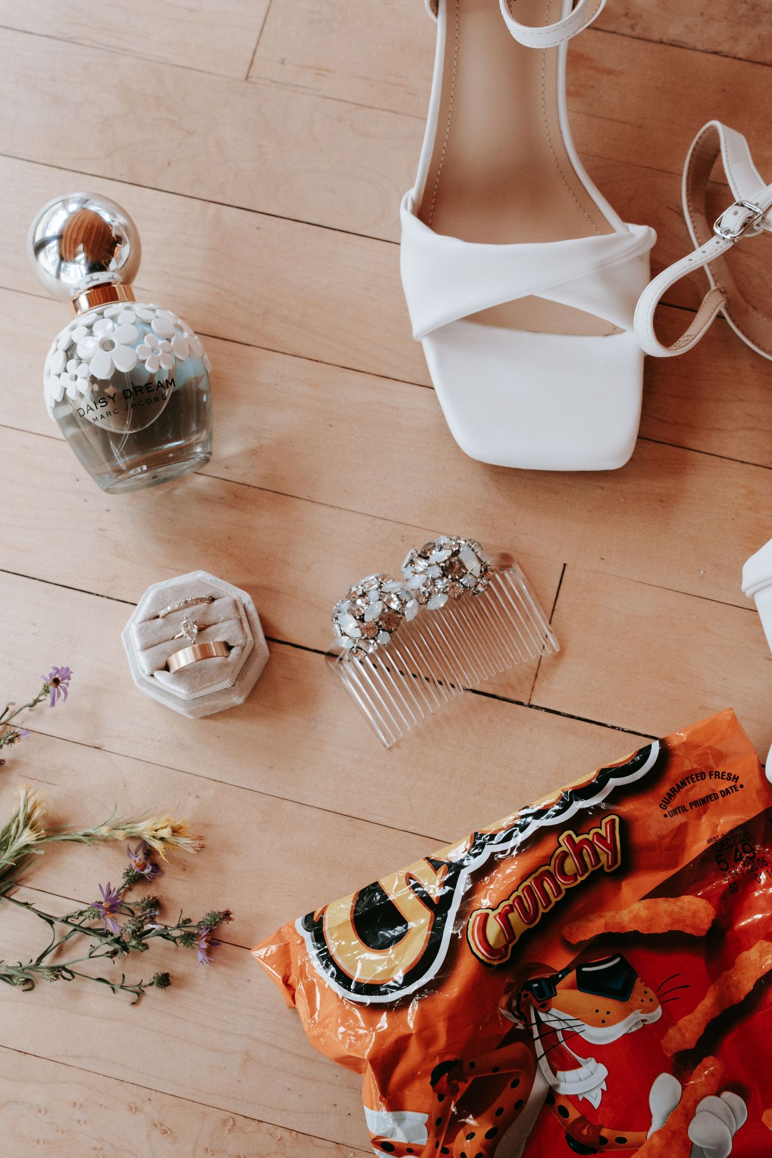 Bridal flat lay featuring white heels, rhinestone hair comb, ring box, perfume, wildflowers, and Crunchy Cheetos on wood floor.