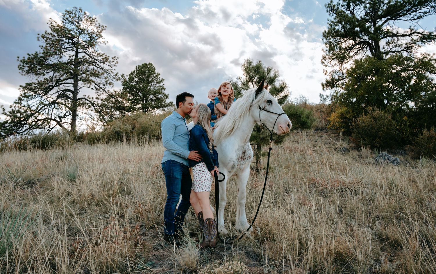 Couple sharing a tender moment beside a white horse in a golden grass field at sunset.