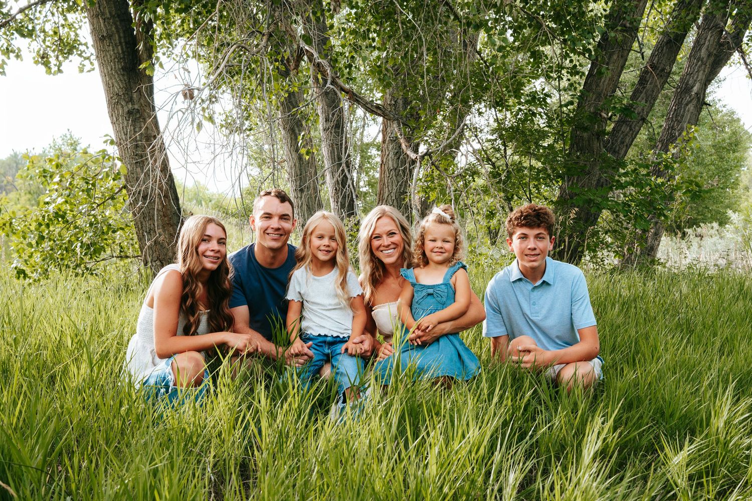 Happy family of six sitting in lush green grass under large trees outdoors.