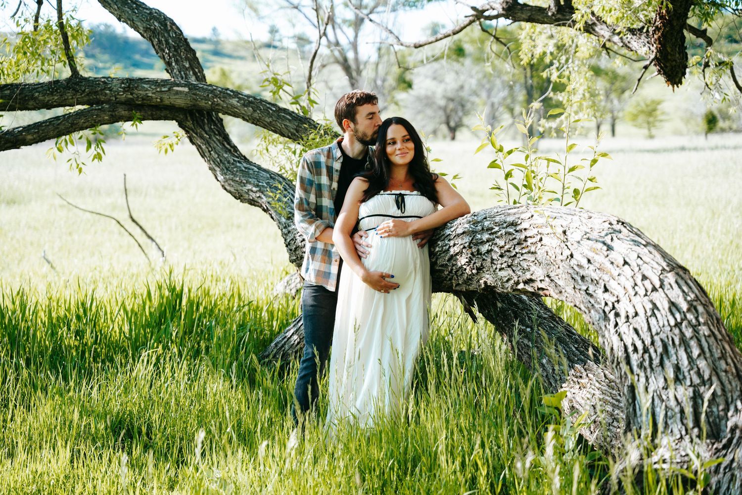 Pregnant woman in white maternity gown embraced by partner while leaning against tree in green field.