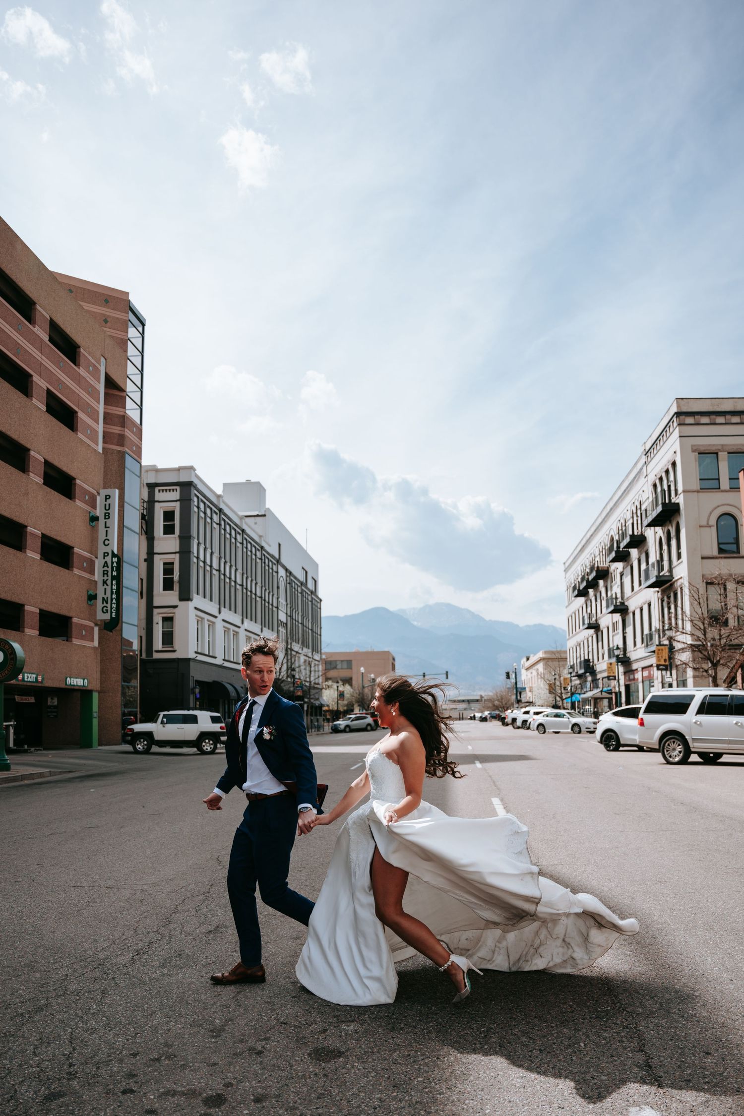 Bride and groom dancing joyfully in urban street with mountain backdrop on wedding day.