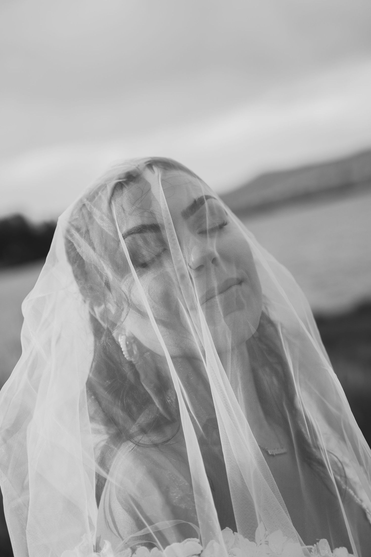 Black and white portrait of a bride with eyes closed, draped in a sheer wedding veil outdoors.