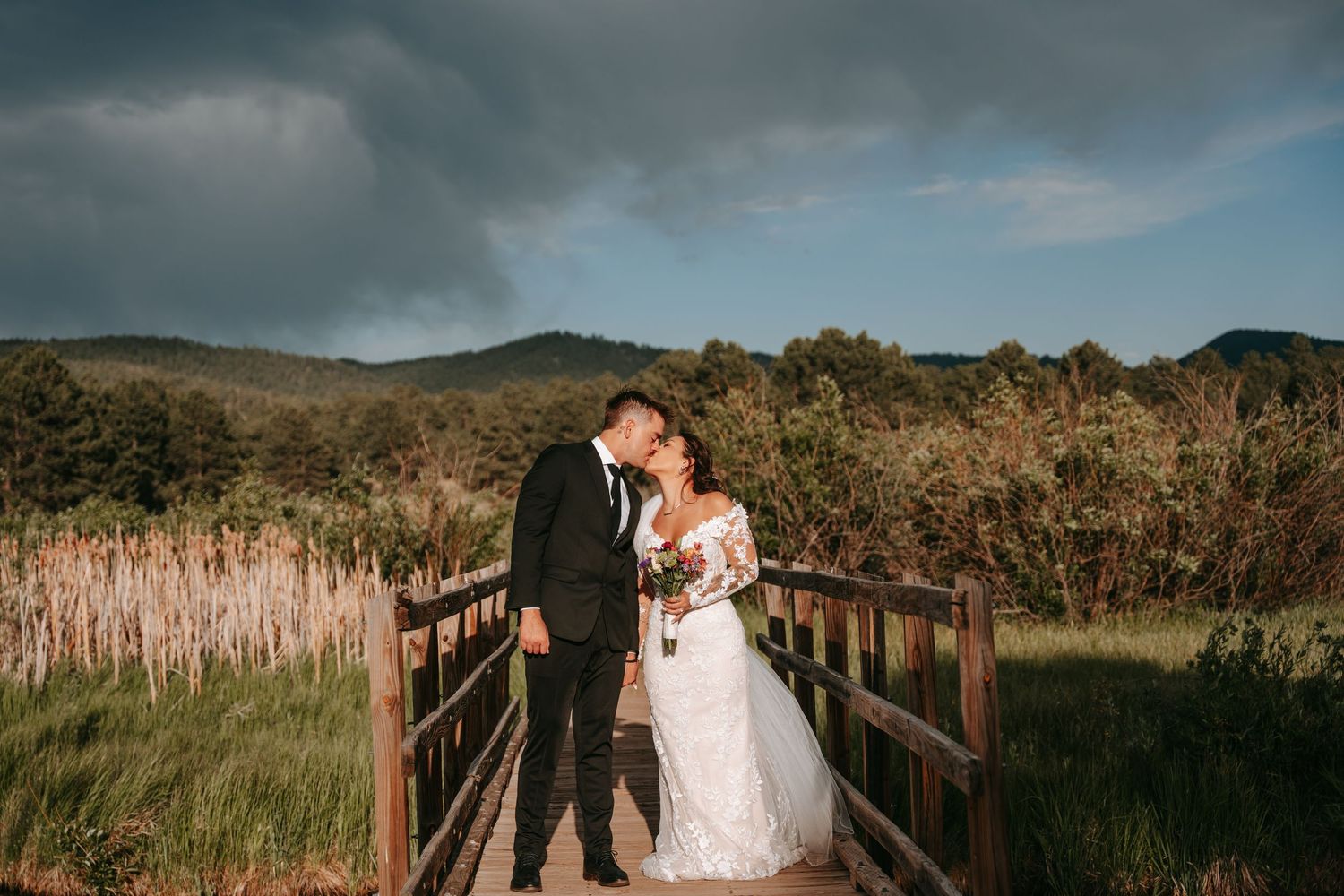 A bride and groom share a romantic kiss on a wooden bridge surrounded by lush greenery and dramatic stormy skies.