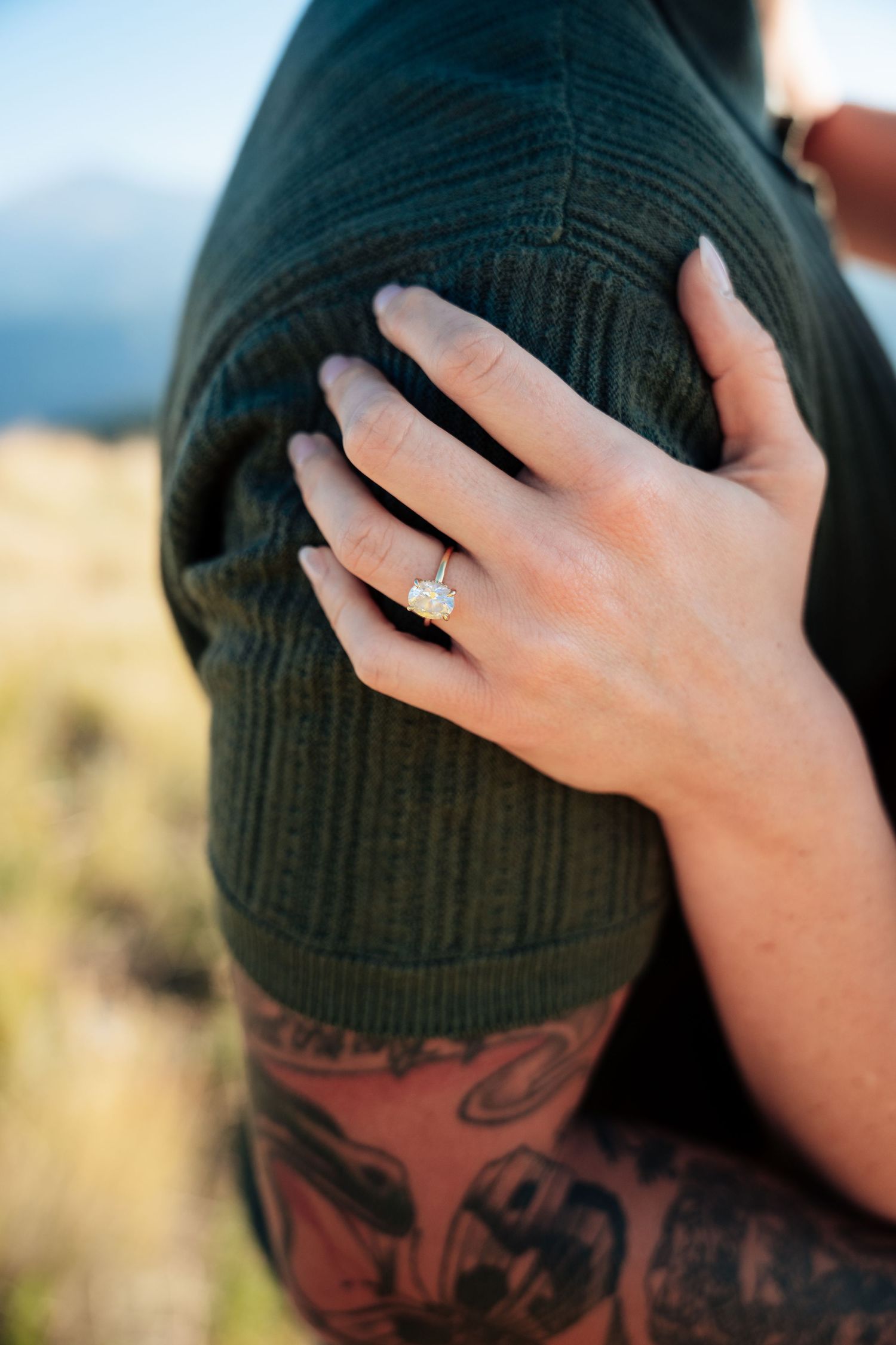 Close-up of a woman's hand with a stunning oval diamond engagement ring resting on her partner's tattooed arm.