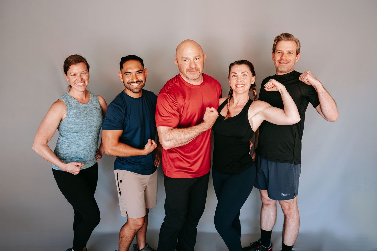 Five smiling fitness professionals flexing muscles together against gray studio background.