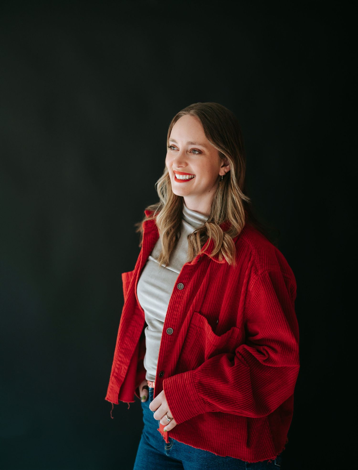 Confident young woman smiling in a red corduroy jacket and white turtleneck against dark background.