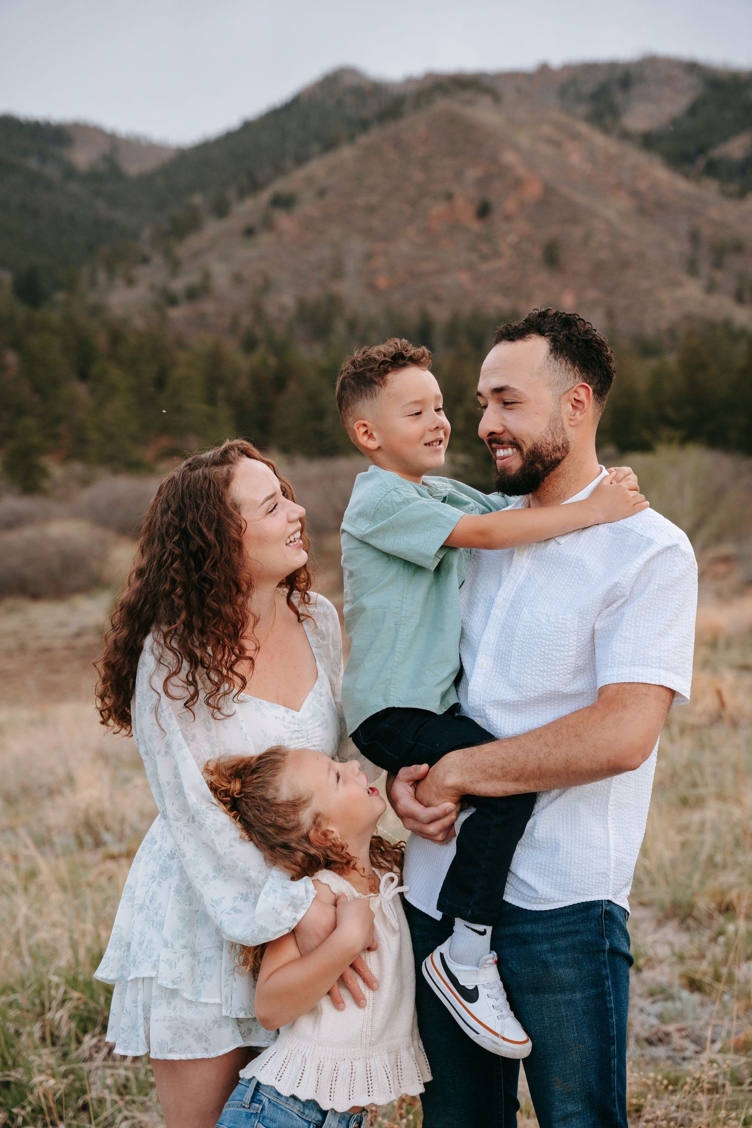 A happy family of four poses outdoors with mountain scenery, parents holding two young children in a warm embrace.